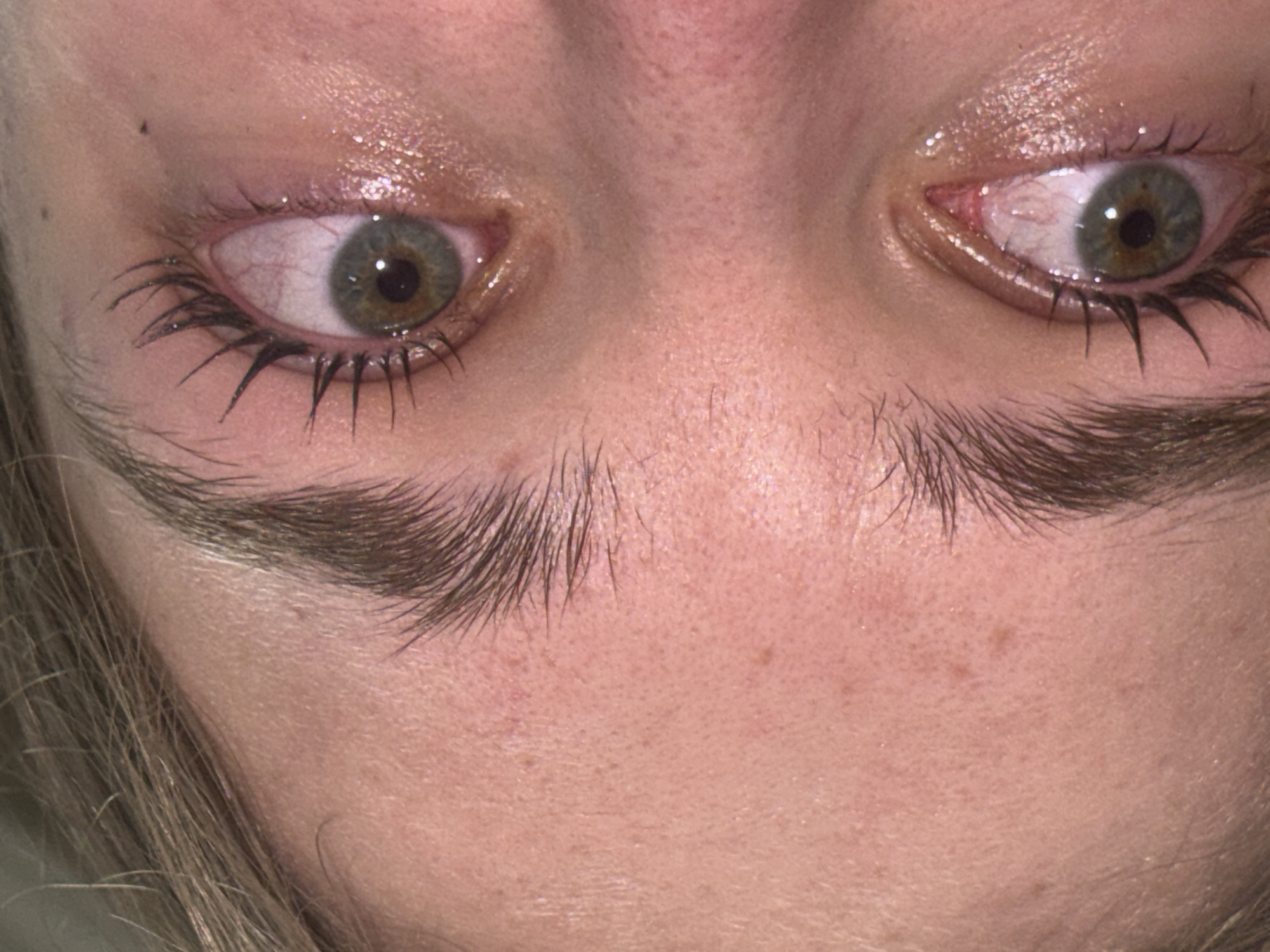 Close-up image of a person's face, focusing on the eyes and eyebrows. The eyes are green/hazel, with visible blood vessels. The eyelashes are thick with mascara. The eyebrows are thick and meet in the middle. The skin tone is fair, with visible pores and some minor freckles. There is no indication of this image being in Perm, Russia.