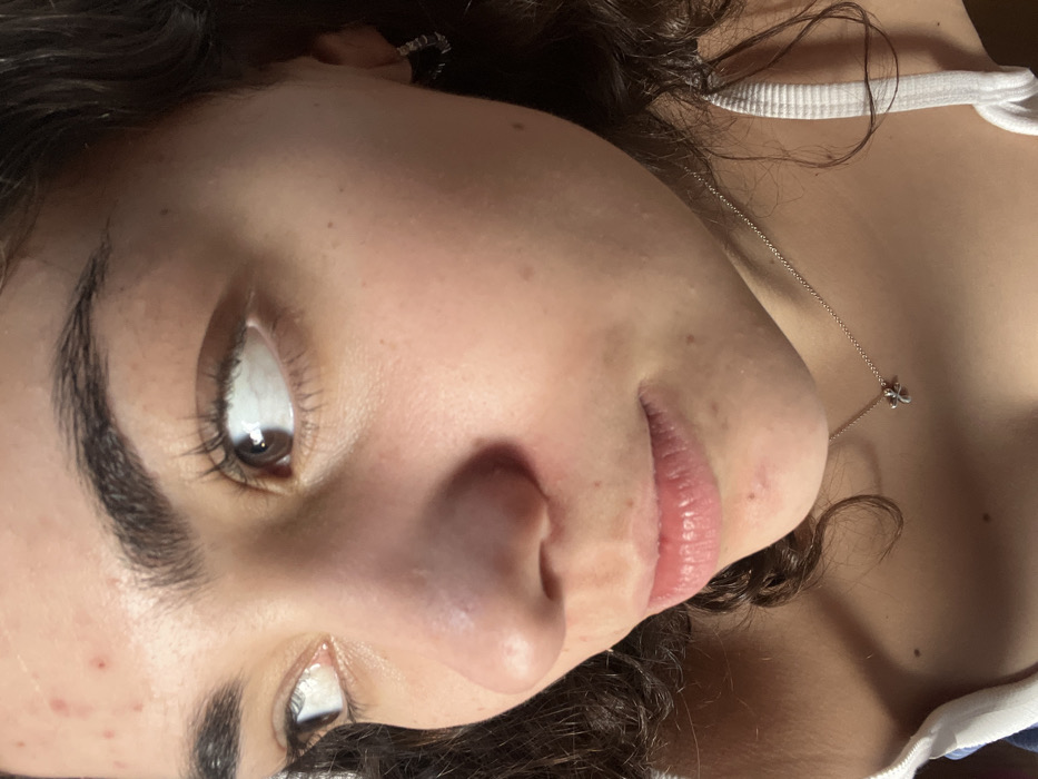 A close-up portrait shows a young woman with fair skin, brown eyes, and dark, curly hair. She wears a white tank top and a delicate silver necklace with a clover pendant. The camera captures her face from a low angle, emphasizing her nose, lips, and thick eyebrows. Her skin has a few small blemishes. She's wearing earrings, in her left ear. The lighting highlights the sheen on her lips and the details in her eyes.