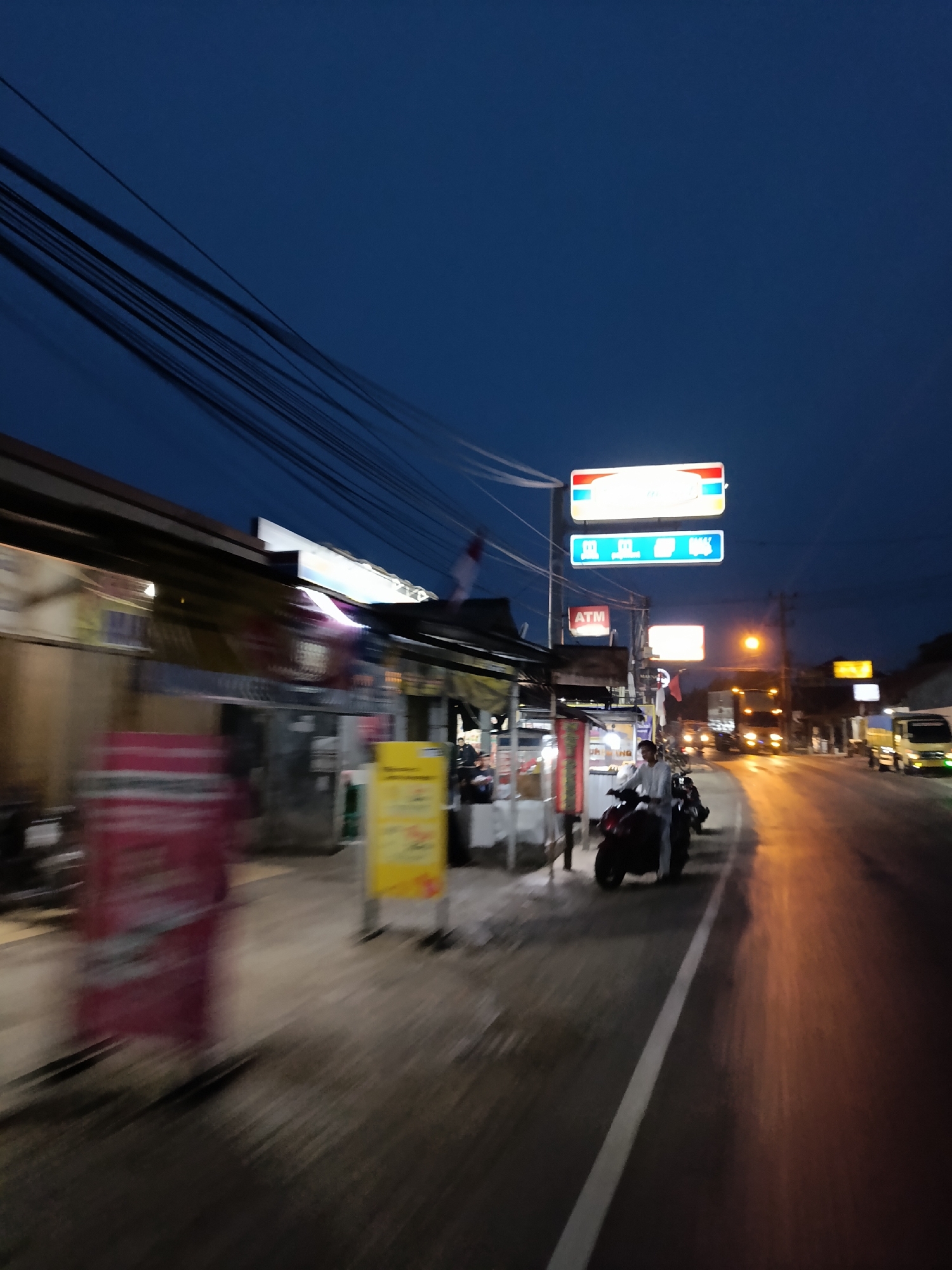 This is a motion-blurred, low-light photograph taken at dusk on a street in Jonggol, Indonesia. The scene depicts a roadside commercial area with several shops and signs. Prominent signs include a rectangular blue sign with white text and a smaller red and white sign with the text "ATM". Overhead, a tangle of power lines crisscross the deep blue twilight sky. In the foreground, a person is riding a dark-colored motorcycle on the right side of the road, which is illuminated by artificial lights reflecting off its wet surface. To the left of the road, storefronts with awnings are visible, with some blurred signage and what appear to be promotional displays. The overall impression is of a busy, active street scene at the transition from day to night.