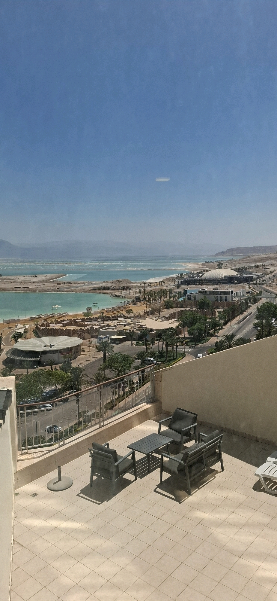 This is a high-angle, vertical, full shot of a resort area on the Dead Sea in Tamar Regional Council, Israel, on a sunny day. The foreground features a tiled balcony with a light beige railing. On the balcony, there is a dark gray outdoor furniture set consisting of two armchairs, a loveseat, and a rectangular coffee table. To the left of the furniture set, a silver umbrella stand is visible. In the mid-ground, a road runs alongside a coastline, lined with palm trees. Beyond the road and trees is the Dead Sea, with its distinctive turquoise water and light-colored salt formations. The background shows a hazy, mountainous landscape under a clear blue sky with a single elongated cloud. The resort area appears to be under development or construction, with several buildings and structures visible.