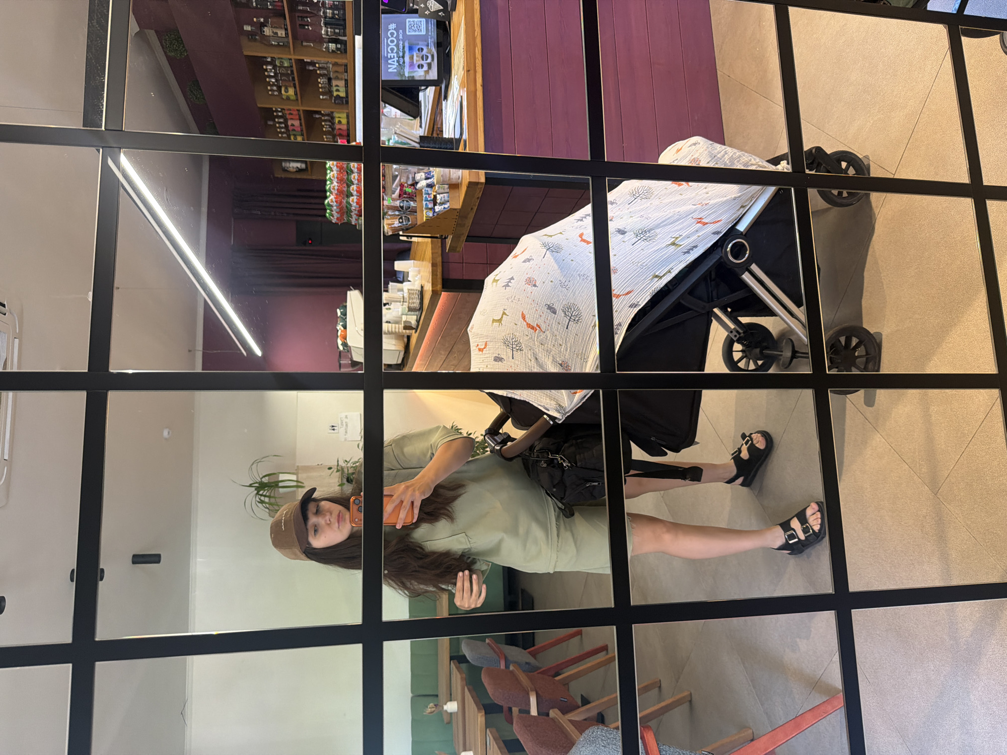 A woman is taking a mirror selfie in what appears to be a cafe or restaurant. She is wearing a brown baseball cap, a light green t-shirt, and light green shorts. She is holding an orange smartphone in her right hand, which is pointed at the mirror. In the background, there is a stroller with a patterned cover. The environment includes tables and chairs, shelves stocked with items, and a counter with a computer monitor. The reflection in the mirror shows the interior of the establishment and the woman's reflection.