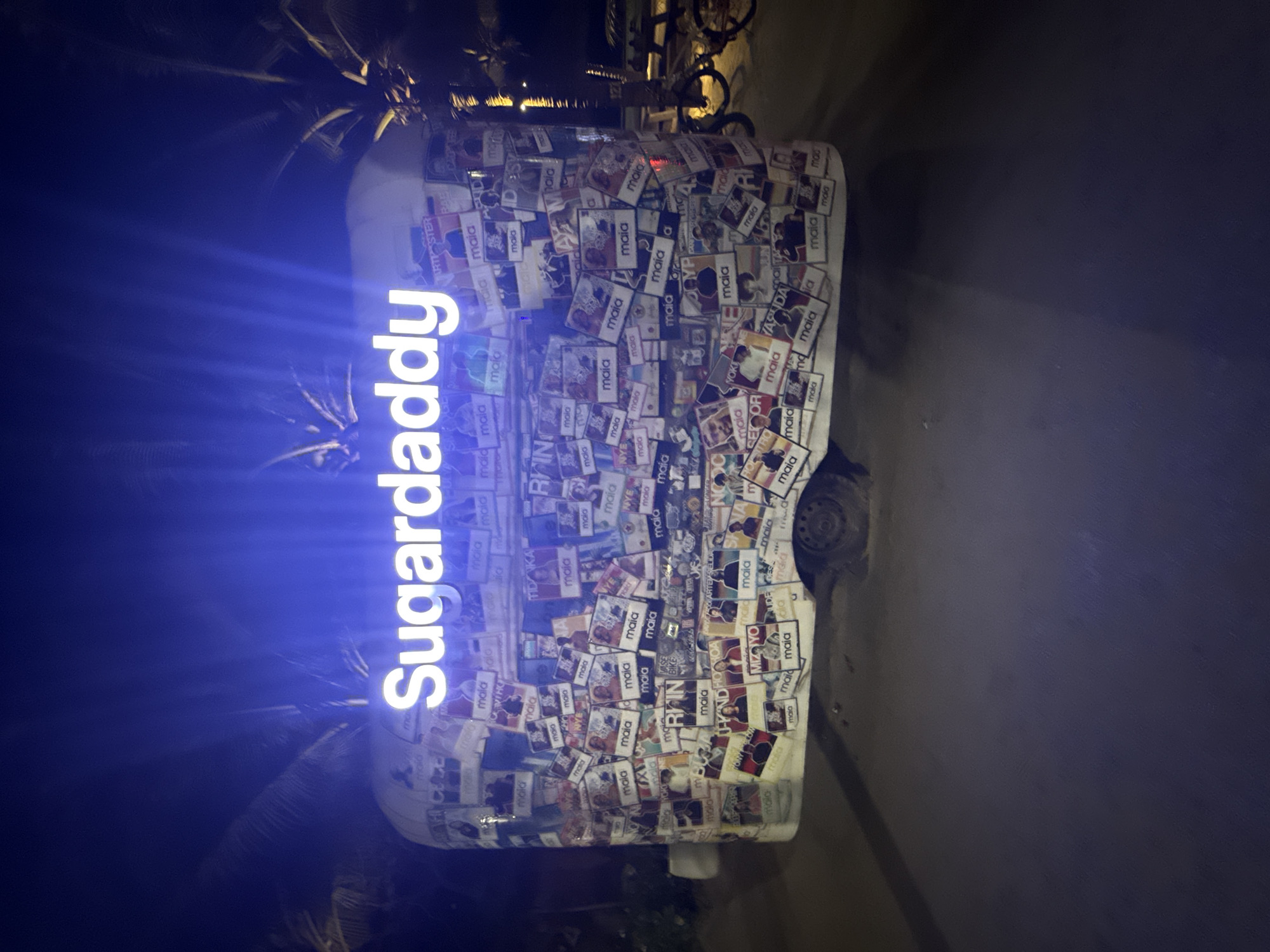 The image depicts a vehicle, possibly a van, covered in stickers, predominantly featuring the word "maia" and other designs. A brightly lit sign reading "Sugardaddy" is visible on the vehicle, casting a blue glow. The setting appears to be outdoors at night, with palm trees visible in the background. The location might be in Đà Nẵng, Vietnam (based on the provided instructions, even though there's no evidence in the image). The surrounding environment is dimly lit. The vehicle seems to be parked on a road or paved area. There are no people explicitly visible in the shot. The stickers and sign are the primary subjects of the image.