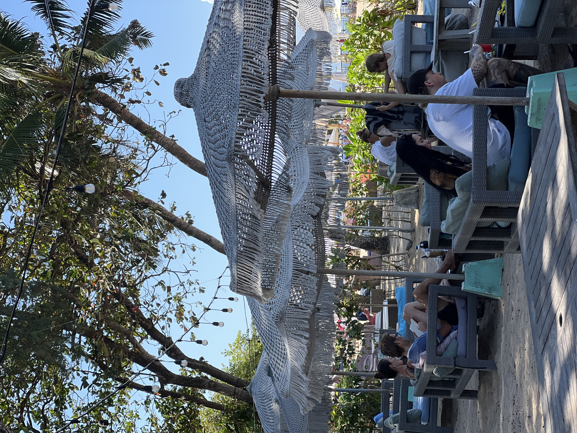 This image captures a vibrant beachside scene in Denpasar, Indonesia, likely at a popular resort or cafe. The foreground features a wooden table with a circular cutout, hinting at a relaxed dining or lounging area. Beyond this, a series of outdoor seating arrangements are visible, composed of grey wicker-style furniture with blue cushions, set upon sandy ground.
Several large, woven umbrellas with tasseled fringes provide shade over these seating areas. The umbrellas are a prominent feature, their intricate texture and bohemian style adding to the atmosphere. In the mid-ground, people are seen relaxing on the lounge furniture. A couple in the left-mid ground are seated, with one person reading a book. Further to the right, another group is gathered, with one individual wearing a black cap and another with long dark hair.
In the background, more beachgoers and umbrellas can be spotted, with the gentle blue of the ocean visible in the distance. Lush green foliage and palm trees frame the scene, interspersed with strings of fairy lights, suggesting the area is enjoyed both during the day and into the evening. The overall impression is one of leisure and tropical ambiance.