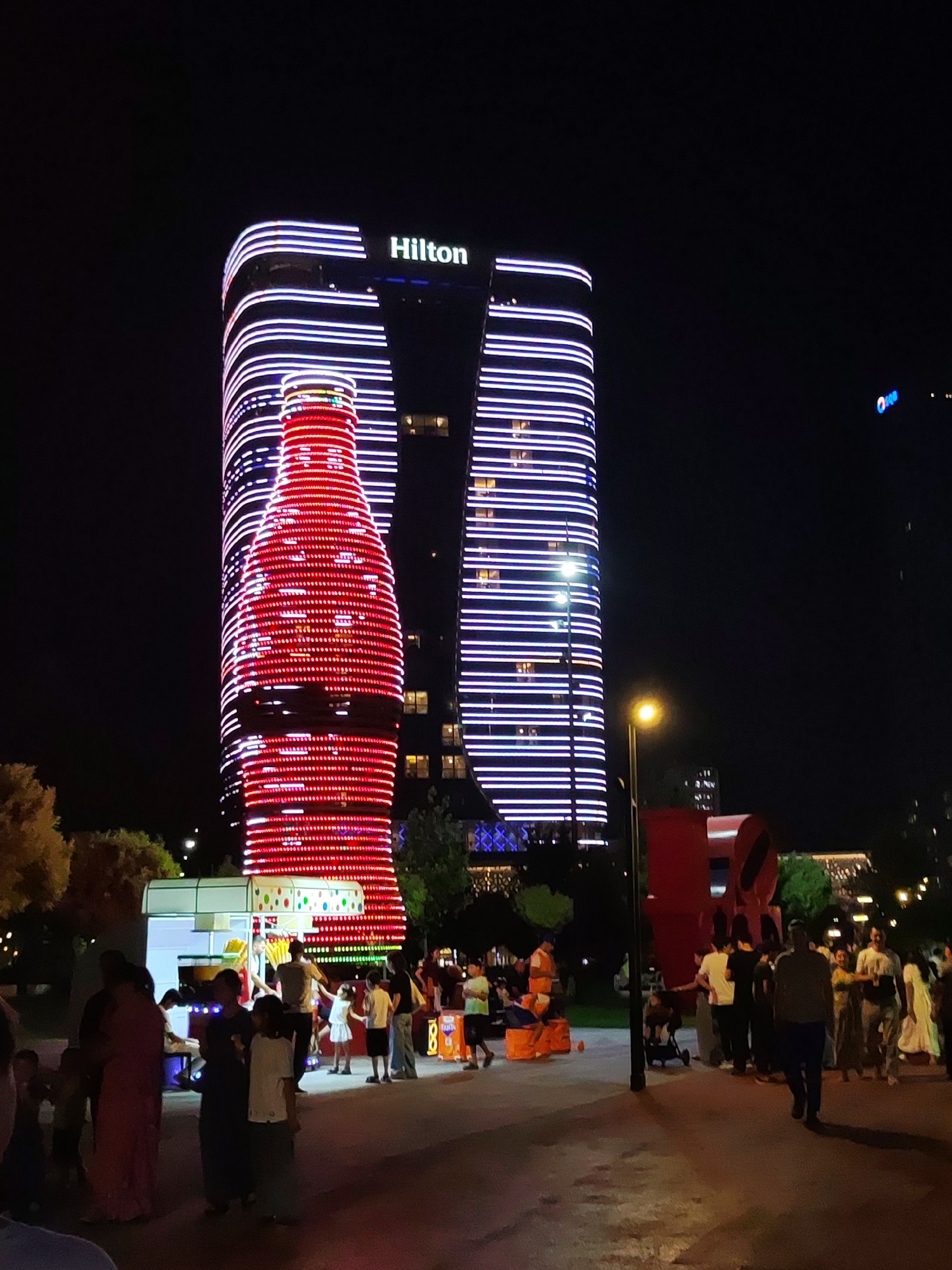 The image depicts a lively night scene in Tashkent, Uzbekistan, featuring the illuminated Hilton Tashkent City hotel. The hotel's facade is a prominent element, with horizontal lines of purple LED lights accentuating its modern architecture. A striking feature is the building's cladding designed to resemble a giant, illuminated red soda bottle, covered in a dense pattern of red LED lights. In the foreground, a crowd of people, including families and children, are gathered, suggesting a public event or gathering. There are various stalls set up, one appearing to be a food or drink vendor with brightly colored decorations. The ground is paved, and streetlights and ambient lighting from the surrounding buildings illuminate the area. The overall atmosphere is festive and energetic, characteristic of a night out in a bustling urban environment.