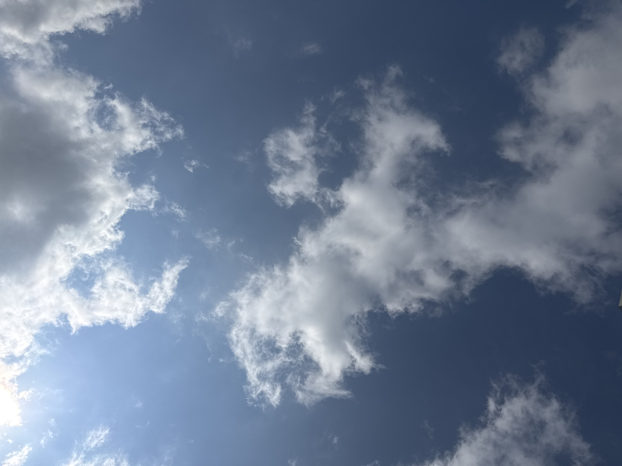 The image captures a bright, cloudy sky over what appears to be a suburban or rural setting, potentially in Nikolaevka, Russia. The sun is partially obscured by dense, white clouds, creating a diffused light. The sky is a deep blue, transitioning to lighter shades where the sun's rays penetrate. Wispy cirrus clouds are scattered across the frame, interspersed with larger, more voluminous cumulus clouds. The clouds exhibit varying degrees of opacity, with some allowing sunlight to pass through, creating a luminous effect. The bottom portion of the image shows a sliver of a building, suggesting an architectural element is present in the scene. The overall impression is one of a pleasant, overcast day, with the clouds dominating the visual field.