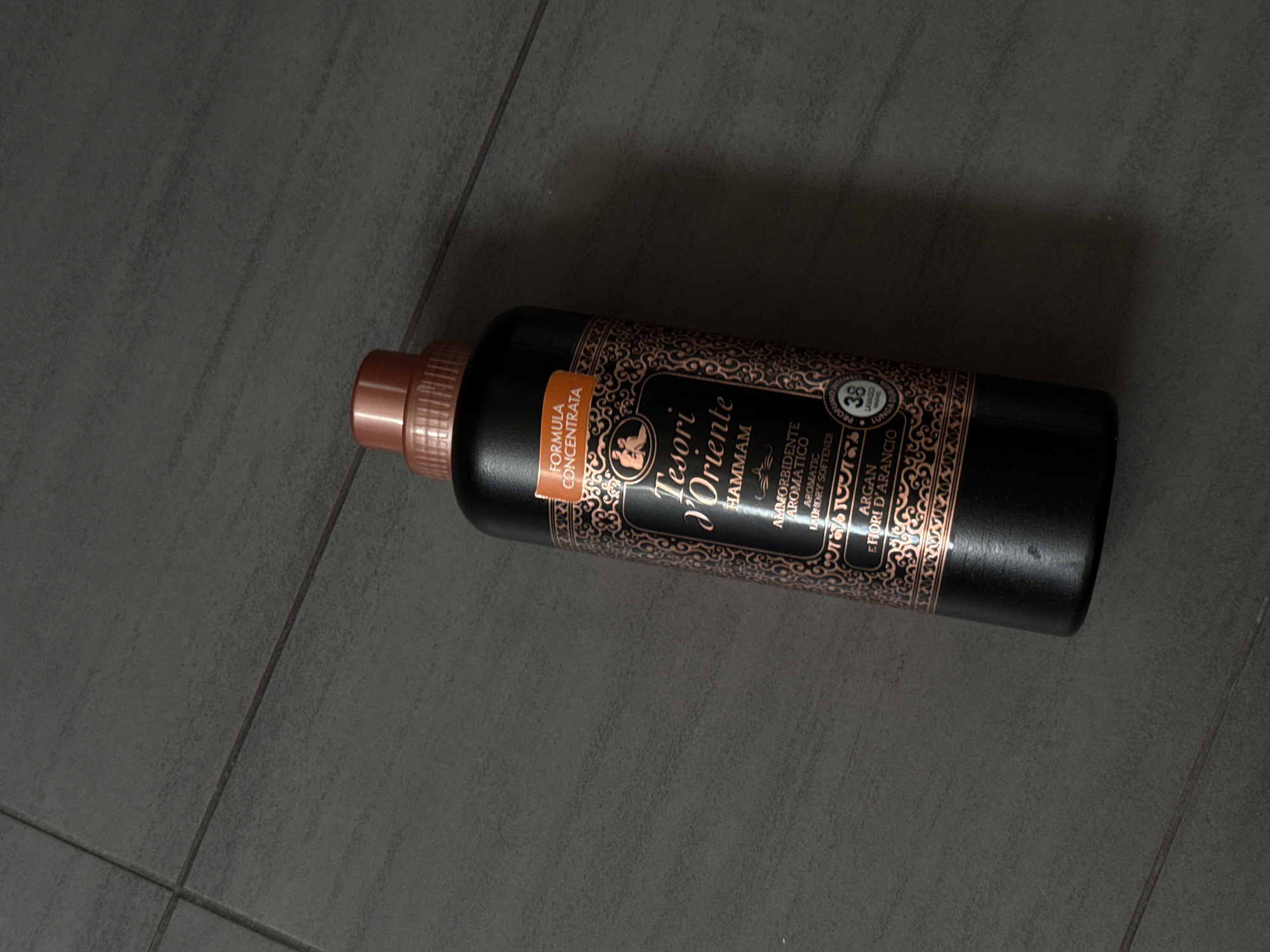 An overhead shot shows a black bottle of Tesori d'Oriente Hammam laundry softener with a copper-colored cap, resting on a dark gray tiled floor. The bottle is oriented diagonally from the bottom left to the top right of the frame. The label on the bottle is predominantly black with intricate copper-colored designs and text. Visible text includes "FORMULA CONCENTRATA," "Tesori d'Oriente," "HAMMAM," "AMMORBIDENTE AROMATICO," "LAUNDRY SOFTENER," "ARGAN E FIORI D'ARANCIO," and a circular emblem with "38 LAVAGGI." The background is a dark gray tiled floor with visible grout lines running horizontally and vertically. The scene appears to be indoors, in a domestic setting. There are no people or animals present. The lighting is even, casting no distinct shadows. The image does not contain any information that would indicate its location within the city of Łódź, Poland.