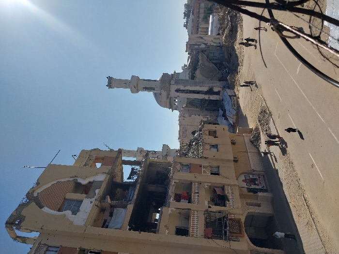 The image depicts a street in Khan Yunis, Palestinian Territory, under a clear, bright blue sky with direct sunlight from the upper right. On the left, a multi-story, light-colored building, likely residential, exhibits severe structural damage; large sections of its facade and interior walls are destroyed, exposing multiple floors, rebar, and internal debris. Laundry is visible hanging from several damaged balconies. An ornate, damaged archway is present on an upper floor. In the background, a large mosque structure is extensively damaged, with its main dome largely collapsed into a substantial pile of rubble. One minaret remains partially standing but shows significant damage.
The paved street runs through the mid-ground, featuring two lanes with faded white line markings. Piles of construction debris are scattered on the unpaved ground adjacent to the road. Approximately 15-20 individuals, dressed in civilian clothing, are observed on the street and sidewalks. A group of five people stands on the left sidewalk. A lone individual walks in the left lane. Further down the street, closer to the mosque, a cluster of approximately six people is visible, one of whom appears to be riding a horse. Additional scattered individuals or small groups are present further along the road. The immediate right foreground is partially obscured by a dark, ornate metal railing.