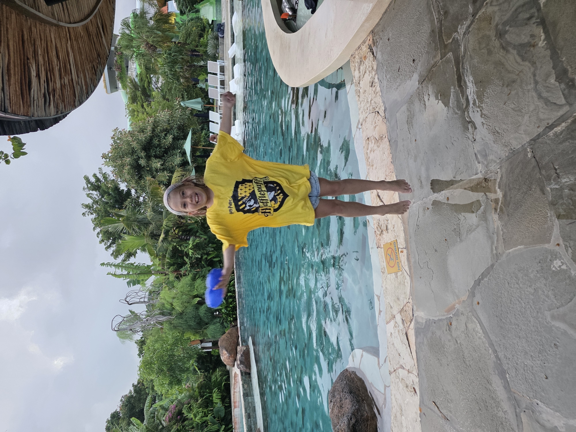 A girl in a yellow Harry Potter Hufflepuff t-shirt stands on the edge of a swimming pool at the Hard Rock Hotel in Kuta, Indonesia. She is smiling and holding a blue toy, with her arms raised slightly. The pool water is clear and turquoise. The pool edge is made of gray stone tiles, with a "No Diving" sign posted on the wall. Lush tropical vegetation and a thatched roof structure surround the pool area. White lounge chairs are visible in the background.