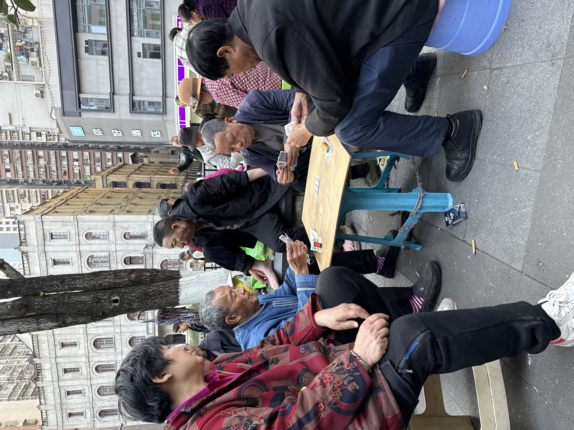In Yuzhong District, China, several older adults are gathered outdoors. Some are seated around a small, light-colored table playing cards. Others stand or sit nearby, observing. The players are primarily men, wearing casual clothing. Cigarette butts litter the ground near their feet. The setting is urban, with buildings of varying architectural styles visible in the background. A tree trunk with a partially painted white base is prominent on the left side of the image. The overall impression is a casual social gathering of elderly individuals in a public space.