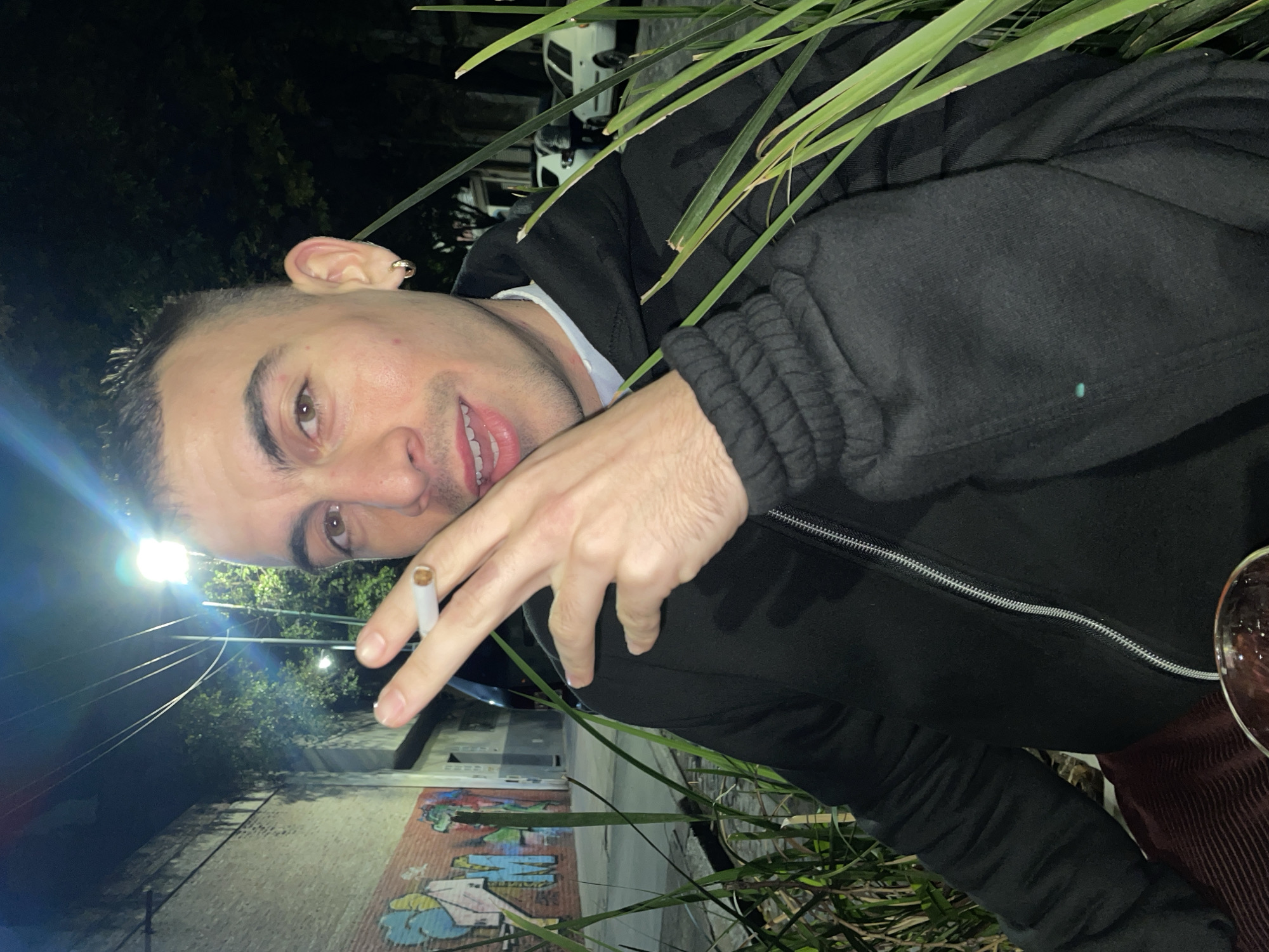 A man is holding a lit cigarette in the fingers of his left hand, with the illuminated end of the cigarette facing to the left. He is angled slightly toward the viewer. He has short dark hair and an earring in his left ear. He's wearing a black jacket, zipped up to his neck, and a light-colored shirt underneath. Green leaves from a plant are visible in the upper right and along the bottom of the frame. In the background, there is a brick wall with graffiti art on it. Streetlights can be seen. A white car is also visible in the background. It is night. The scene appears to be located in the city of Buenos Aires, Argentina.