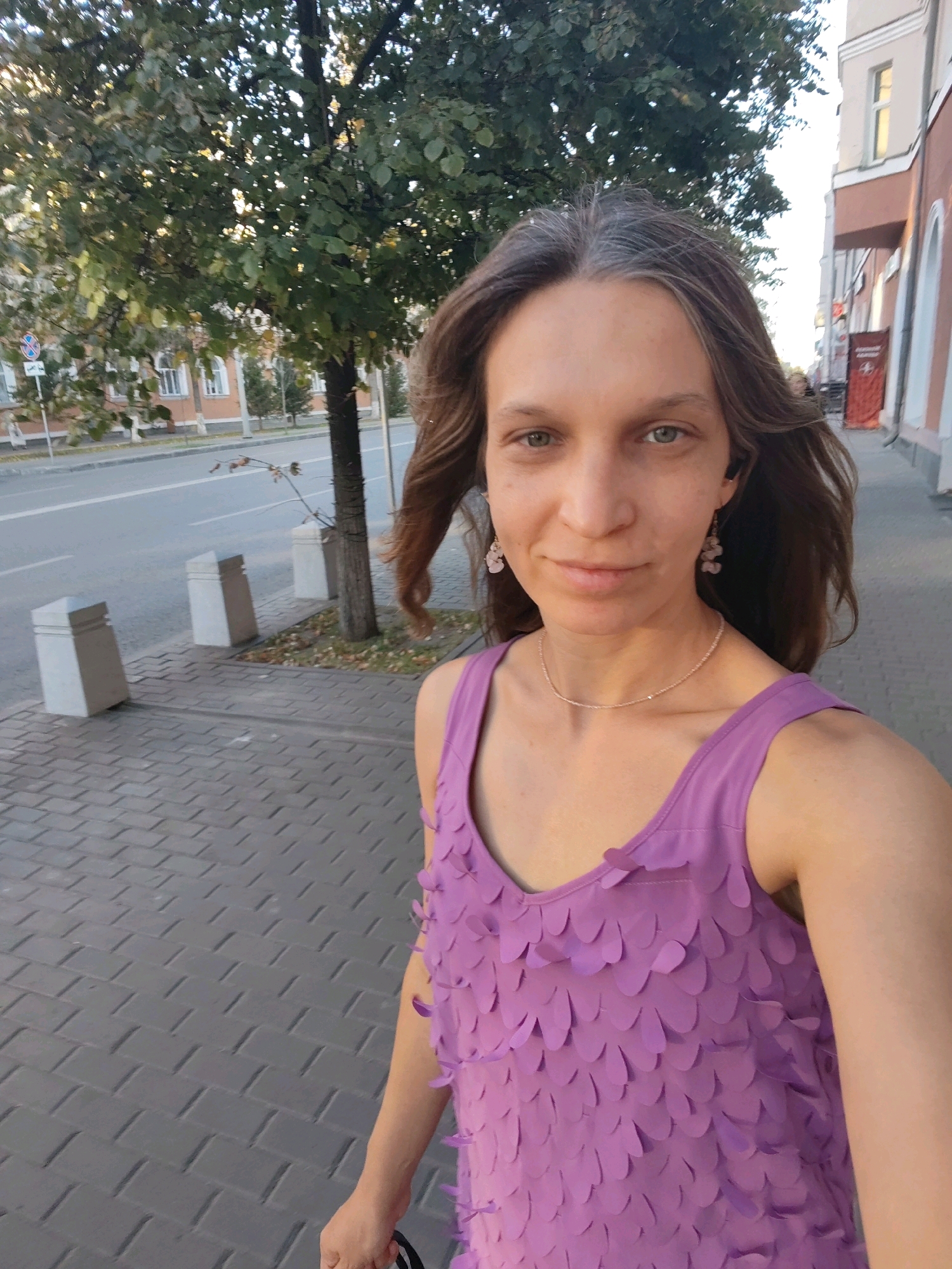 A woman with long, wavy brown hair and green eyes is taking a selfie on a sidewalk. She is wearing a purple sleeveless dress with petal-like embellishments, a delicate necklace, and pink earrings. She has dark earbuds in her ears. The sidewalk is paved with grey rectangular stones and bordered by a tree on the left and buildings on the right. A street is visible behind the tree, with cars and buildings in the distance. The lighting suggests it is late afternoon or early evening.