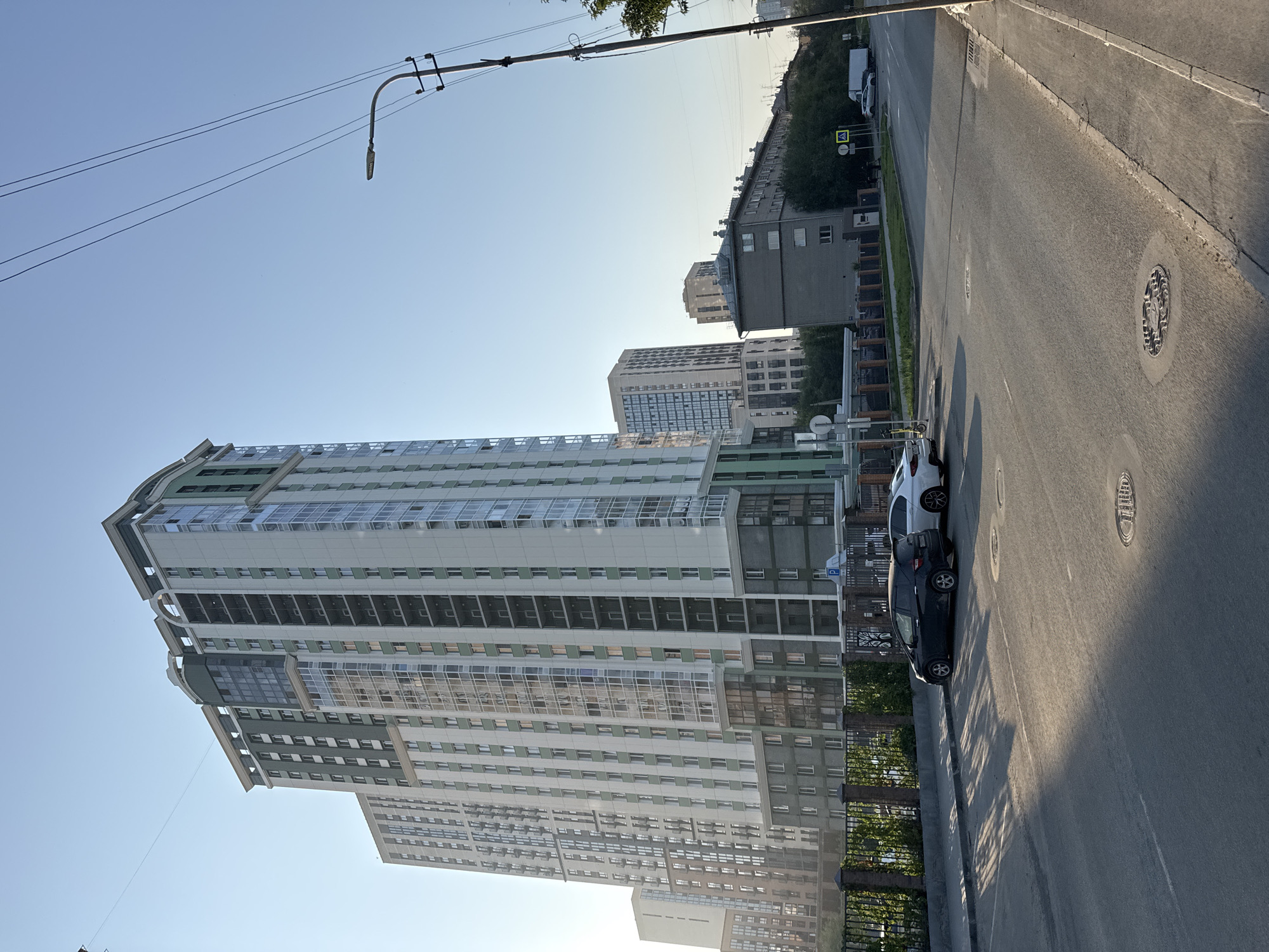 The image depicts a street scene in Novosibirsk, Russia, likely in a residential or mixed-use urban area. A tall, modern residential building with a light-colored facade and green accents dominates the left side of the frame, featuring numerous balconies and large windows. In the foreground, a two-lane asphalt road is visible, with two cars parked on the right side. A black hatchback is parked closer to the viewer, and a white sedan is parked behind it. A sidewalk runs alongside the road, bordered by a decorative metal fence and greenery. In the background, other buildings of varying heights and architectural styles are visible, including a mid-rise apartment building and a longer, lower-rise structure. Overhead, power lines and a street lamp stretch across the clear blue sky. The lighting suggests it is late afternoon or early morning, with long shadows cast across the road. There are no people visible in the immediate vicinity of the parked cars.