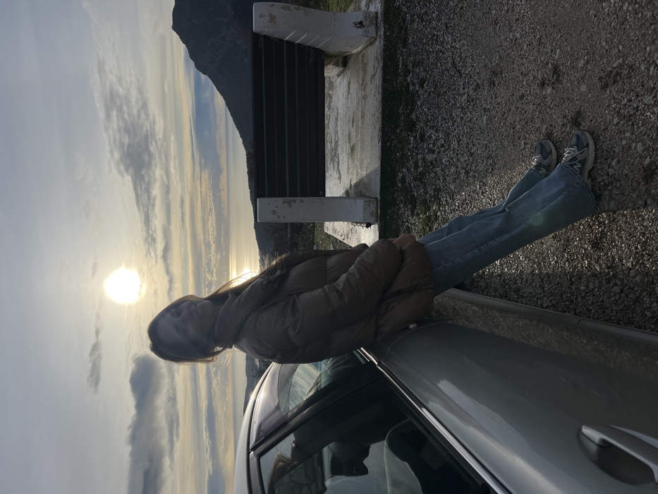 A woman in a brown puffer jacket and blue jeans sits on the hood of a silver car. She faces the sunset over a landscape with a mountain in the background. A concrete bench is visible behind her. The ground is covered in pebbles, and the sky is filled with clouds.