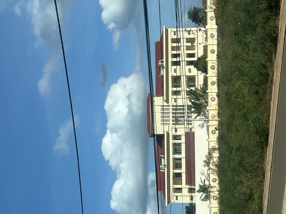 The image displays a large, multi-story building with pale yellow walls and dark red tiled roofs, situated in Trung Hồ, Vietnam, under a bright blue sky with scattered white cumulus clouds. The building features a prominent central section with vertical louvered elements and multiple wings with numerous dark-paned windows. Rooftop equipment, likely ventilation or air conditioning units, is visible on the red roofs. In the mid-ground, a light-colored perimeter wall, possibly concrete or stucco, defines the building's compound. This wall is decorated with regularly spaced circular yellow five-pointed star emblems. Tropical vegetation, including broad-leafed plants and small trees, is visible both behind the wall within the compound and as dense, overgrown green foliage in the immediate foreground. One individual is observed bent over in the overgrown area in front of the decorated wall, slightly to the left of the image center, engaged in an indiscernible ground-level activity. Dark horizontal lines, consistent with power or communication cables, traverse the upper portion of the frame, and a dark grey surface, likely a road or path, is present at the bottom edge.