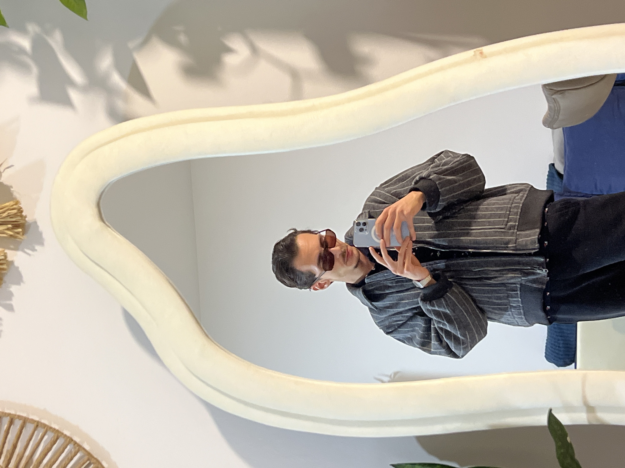 A person is taking a selfie in a mirror. The person is wearing sunglasses, a gray striped jacket, and black pants. The mirror has a thick, off-white frame. In the background are shadows of leaves on a white wall, and a decorative woven object is visible in the lower left corner. The location is not discernible from the image.