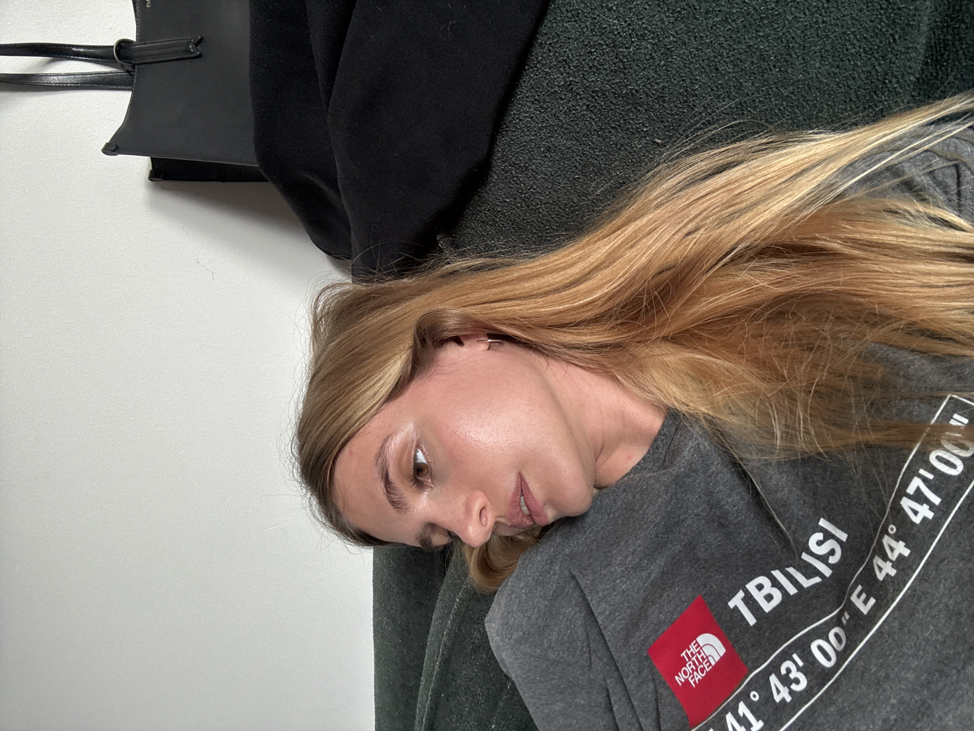 A woman with blonde hair is lying down. She is wearing a gray t-shirt with "THE NORTH FACE" and "TBILISI 41° 43' 00" E 44° 47' 00"" printed on it. A black tote bag is hanging on a white wall above her, and a black jacket is bunched on the couch she is lying on.