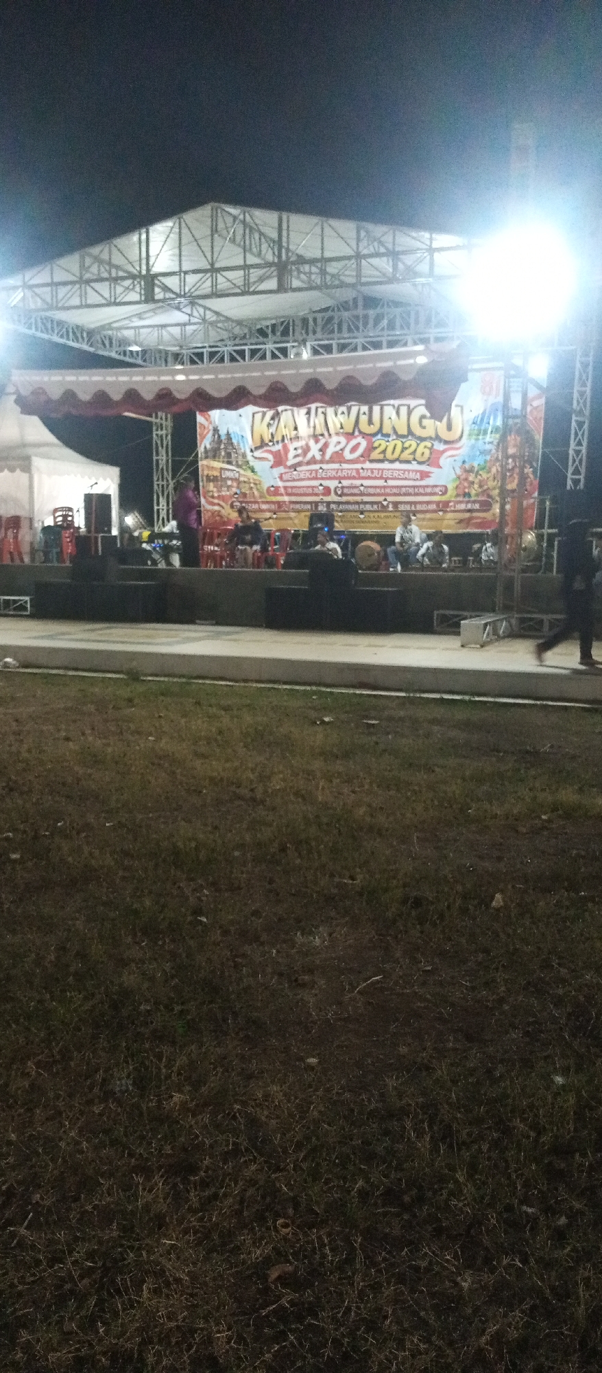 The image depicts an outdoor stage setup at night, likely for an event. A large banner is prominently displayed on the stage, reading "KAWUNG EXPO 2026". The banner features colorful graphics, including what appears to be a temple and various figures. Several people are visible on the stage, some seated and seemingly engaged in a performance, possibly musical, given the presence of drums. A white tent structure is situated to the left of the stage, and lighting equipment is visible, illuminating the scene. In the foreground, a grassy area is visible, and to the far right, a person is walking away from the camera. The overall setting suggests a public event or festival. The city of Kaliwungu, Indonesia, is identified as the location, implying the event is taking place there.