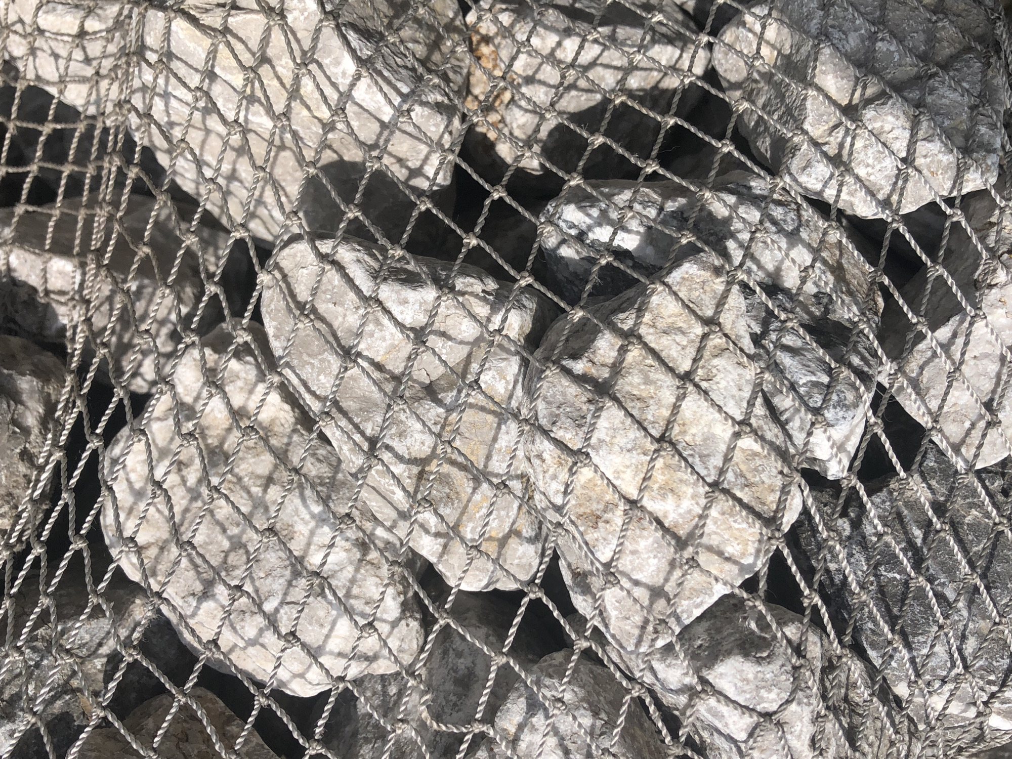 The image shows a close-up, overhead view of a gabion wall, a retaining structure filled with rocks and enclosed by wire mesh. The rocks are of varying sizes and are predominantly light gray and beige, with some darker gray ones visible. The wire mesh is a coarse, woven net with a diamond pattern, creating a textured overlay on the rocks. The lighting suggests it is daytime, casting distinct shadows from the mesh onto the rocks and creating bright highlights on the uneven surfaces of the stones. The scene appears to be outdoors, possibly a construction site or part of landscaping, but there is no specific context to determine its location within the city of Krasny, Russia. There are no people or discernible actions depicted.