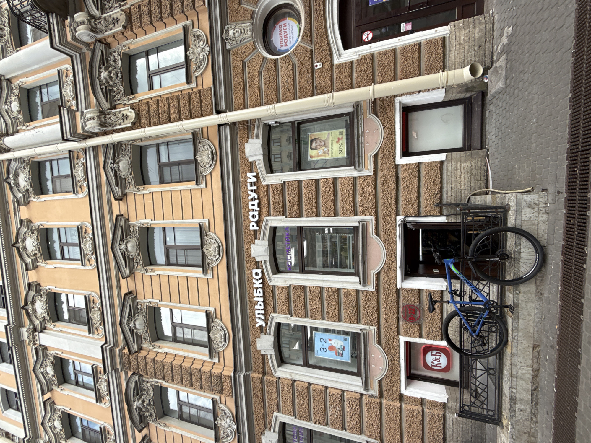 The image displays a multi-story building facade, rotated 90 degrees counter-clockwise, showcasing ornate architectural elements and ground-level shopfronts. The building's walls are a pale orange or beige, interspersed with textured stucco panels and adorned with elaborate greyish-white decorative moldings around windows and between floors. A prominent white drainpipe runs down the center of the facade.
The upper sections feature numerous rectangular windows, each framed by decorative stucco. The ground floor, which appears on the right side of the rotated image, hosts commercial establishments. A circular sign near the top reads "УЛЫБКА РАДУГИ" (Ulybka Radugi, meaning "Smile of the Rainbow"), with additional text "ТОВАРЫ ХИМИИ" (Chemical Goods) and "КОСМЕТИКА" (Cosmetics), indicating a store selling household and beauty products. The store name "УЛЫБКА РАДУГИ" is also vertically inscribed in large letters on the building's exterior.
Several shop windows display posters. One features a person's face and a "-30%" discount notice. Another window shows a poster with "3-2". Towards the bottom, a bright red sign with "К&Б" (K&B) in white letters is visible in a window, accompanied by a small white sign depicting a crossed-out cigarette, denoting a no-smoking area.
A blue mountain bike is suspended from or chained to a black metal railing outside one of the lower windows. To the far right, a dark, textured surface, possibly a sidewalk grate or a rough wall, is visible. The reflections in the windows suggest an overcast daytime, with no direct sunlight. No individuals are present in the scene, which primarily focuses on the building's architecture and commercial signage.