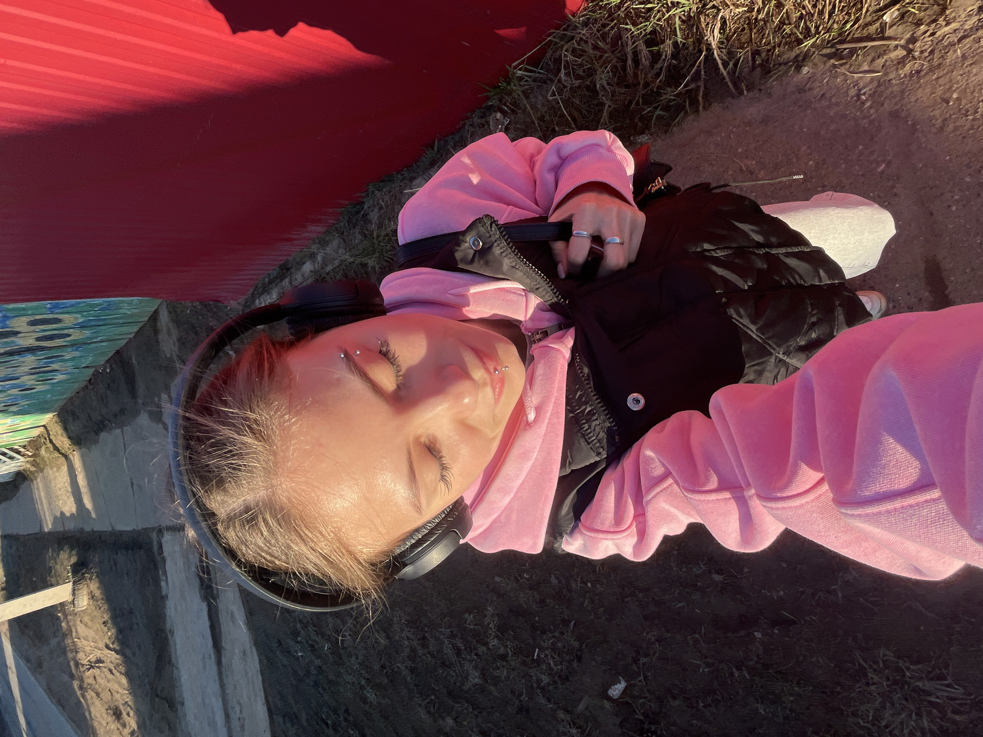 The image captures a high-angle, rotated view of a young person reclining outdoors. The subject, likely female, has light-colored hair and is wearing black over-ear headphones, a bright pink hooded sweatshirt, and a black puffer vest. They have noticeable facial piercings, including an eyebrow piercing and a labret piercing, and wear rings on their fingers. Their eyes are closed, and their face is tilted upwards, basking in direct, warm sunlight, suggesting a moment of relaxation or enjoyment of the sun.
To the upper left of the frame (top in the original orientation), there is a prominent, vibrant red structure, possibly a metal fence or wall, which reflects the strong sunlight. Below this, a section of a colorful, painted surface with blue and green patterns is visible. The immediate ground around the person is covered with dry grass and patches of dirt or a path. The strong shadows and highlights on the subject and environment indicate a bright day, likely morning or late afternoon. No visible text is present in the image.