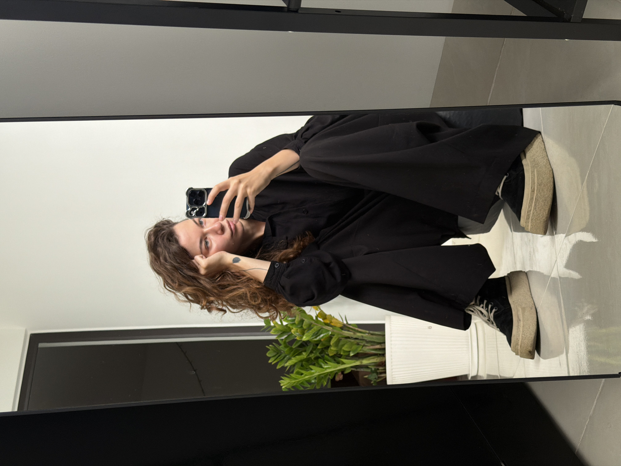 A mirror reflection shows a woman with curly brown hair wearing a black outfit and sneakers. She holds a smartphone up to take a selfie. Beside her, a potted plant sits on a surface that reflects in the mirror. The surroundings include a window and tiled flooring, suggesting an interior setting. The location is not identifiable as Đà Nẵng, Vietnam, based on the visual details provided.