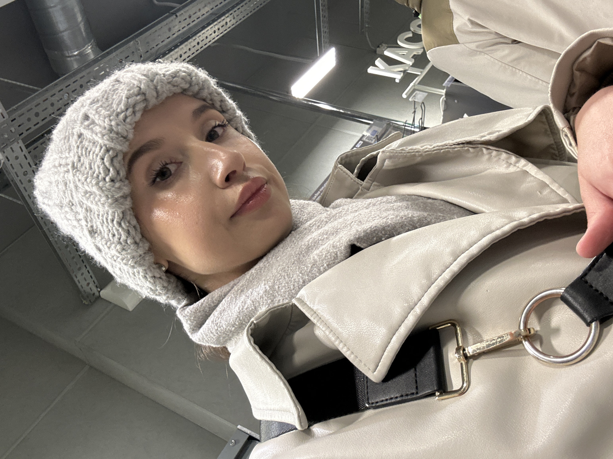 A low-angle, close-up shot captures a young woman looking directly into the camera. She wears a light grey, knitted beanie hat with a textured pattern and a matching light grey scarf or high-collared sweater. Her outer garment is a light beige or off-white jacket, possibly faux leather, featuring a prominent collar and a visible black strap with a silver D-ring and a gold-colored clasp on her right side.
The woman's face is prominent, displaying dark eyes, well-defined eyebrows, and full lips with a pink tint. Her expression is neutral, possibly contemplative, with a subtle pout.
The background reveals an indoor setting with an industrial aesthetic. Exposed metal structures, possibly ventilation ducts or cable trays, are visible on the grey ceiling. Bright rectangular fluorescent lights illuminate the space. In the upper right background, the Cyrillic word "КАССА" (KASSA), meaning "cash register" or "checkout" in Russian, is clearly visible, suggesting the location is a store or commercial establishment in Yekaterinburg, Russia. The overall impression is a casual selfie taken in a public, retail environment.