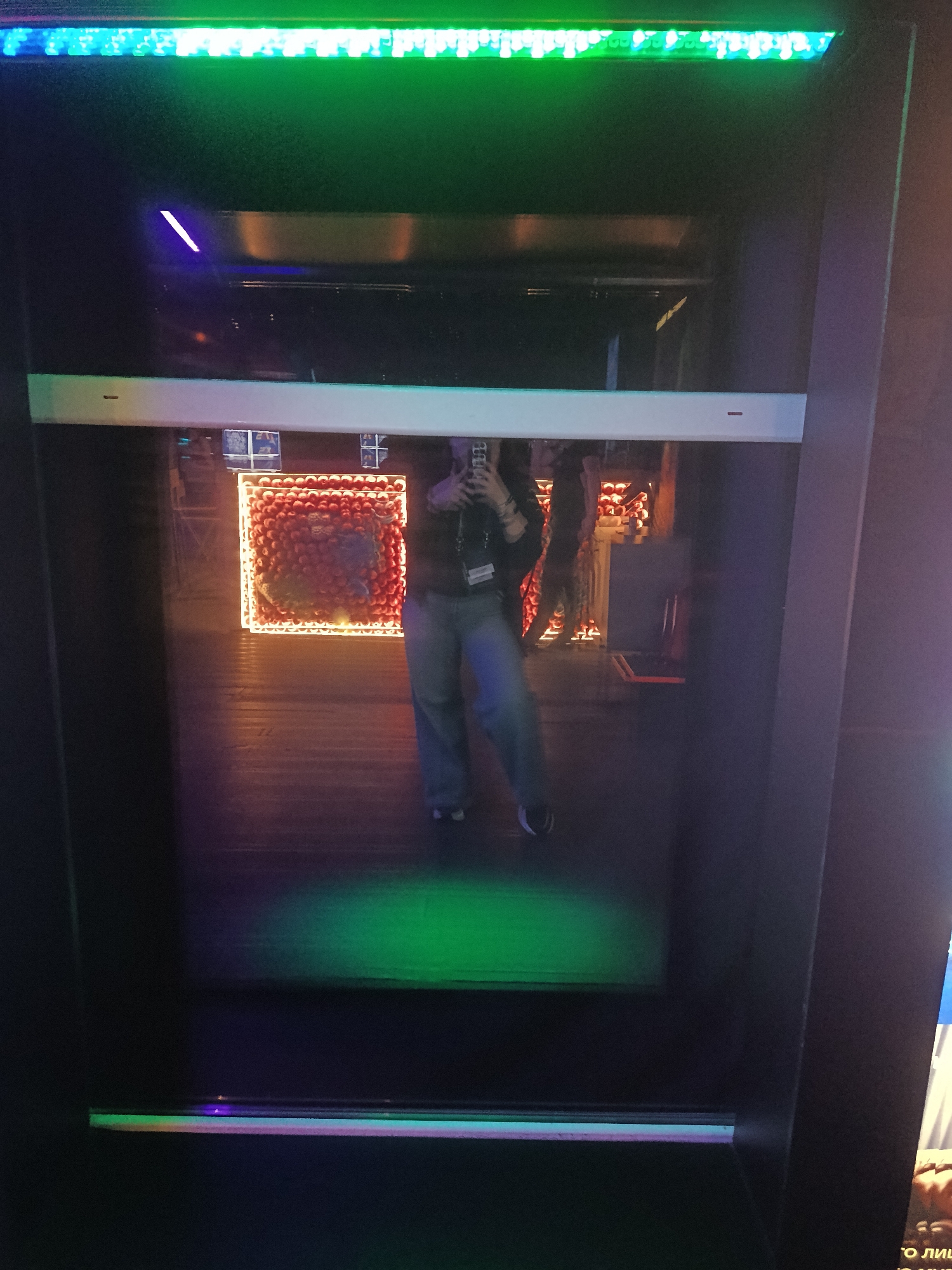 The image shows a reflection in a dark display cabinet or screen, possibly located in Moscow, Russia (based on potential Cyrillic text at the very bottom right corner, not fully visible). The reflection captures a person taking a photo of themselves in the mirror-like surface. They are wearing casual clothes (jeans and a dark top) and have a lanyard around their neck. Behind the person, the reflection reveals a room decorated with a lighted display, possibly a wall decoration made of red orbs surrounded by a glowing frame. Other individuals are also partially visible in the reflection behind the main subject. The cabinet or screen is bordered by dark material, with green and possibly blue lighting along the top and bottom edges. A broad white strip bisects the frame, and there is a green spot-like illumination towards the lower section.