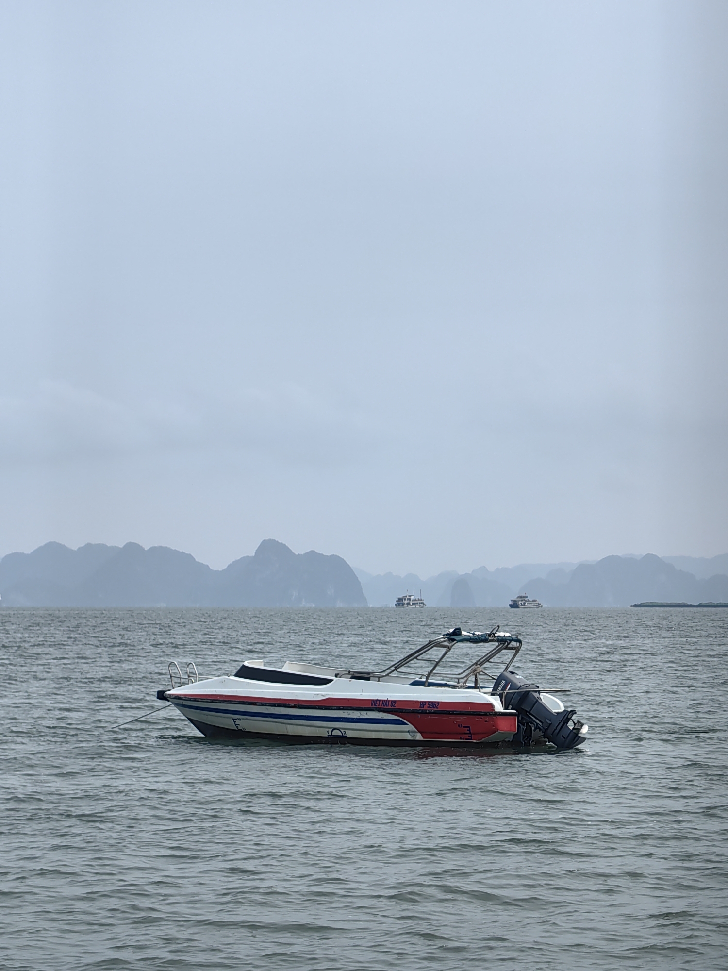 A medium shot depicts a stationary white and red speedboat on choppy, grey water, with distant limestone karsts and overcast sky.
The boat is predominantly white with a thick red stripe along its lower hull and a thinner blue stripe above. The boat's rear has a Yamaha outboard motor attached. Text is visible on the back of the boat, including "VIỆT HẢI 02" and "HP 5952".
In the background, the grey sky blends into distant limestone karst formations, typical of Ha Long Bay. Several ships are visible in the distance. The water is choppy.
This location is in Bai Chay Ward, Vietnam.