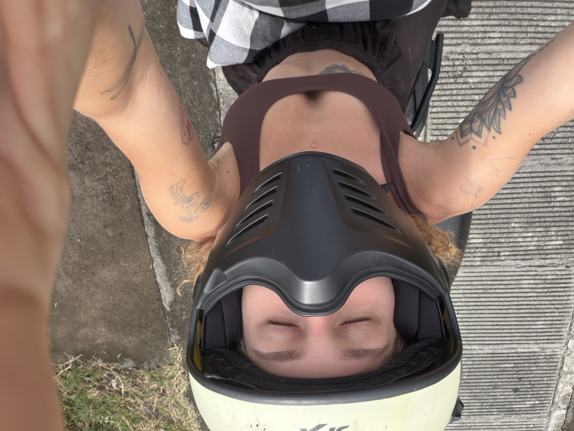 A first-person overhead shot shows a person wearing a brown tank top, a black and white checkered shirt, and a cream and black motorcycle helmet. The individual has their eyes closed. Tattoos are visible on their arms. The location appears to be North Kuta, Indonesia, as indicated by the style of the concrete surface and the presence of tropical vegetation.