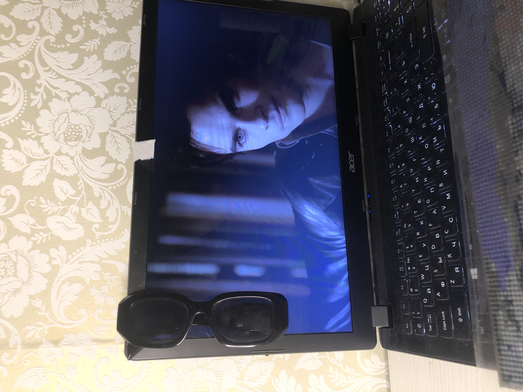 The image shows an open Acer laptop resting on a patterned tablecloth. The laptop screen displays a scene with a person with fair skin and blue eyes, presumably a character from a video or film. A pair of black sunglasses sits at the bottom of the laptop screen, closer to the viewer. The laptop keyboard, with Russian Cyrillic letters, is visible on the right side of the screen. A piece of torn tape is visible on the top-left edge of the laptop screen. The image does not contain information regarding the location in the city of Noviy, Russia.