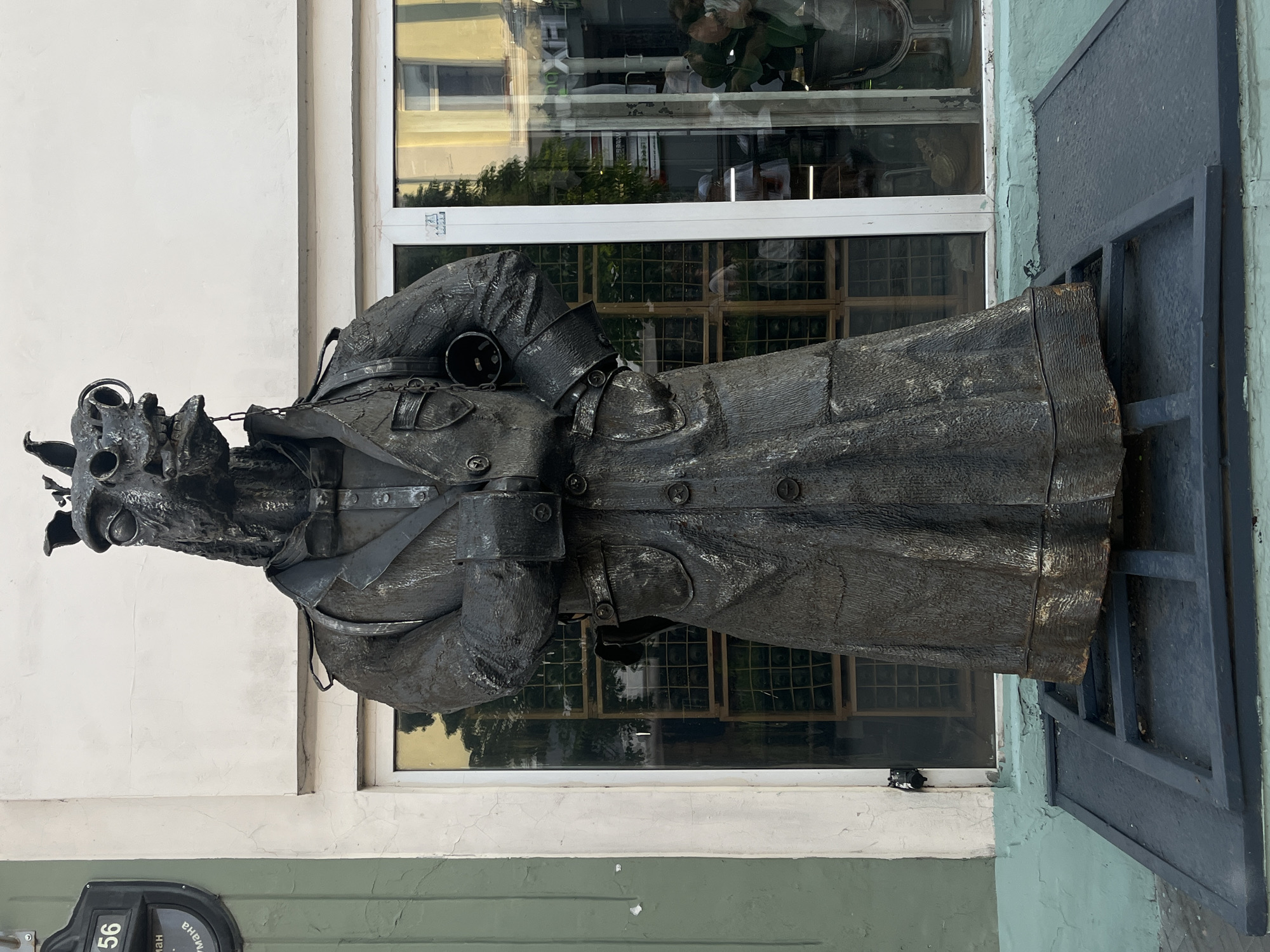 The image shows a metal sculpture of a person in a long coat, likely depicting a historical figure or literary character. The sculpture stands in front of a building with a white facade and large windows. A street sign with the number "56" is visible on the bottom left. The location is Kazan, Russia.