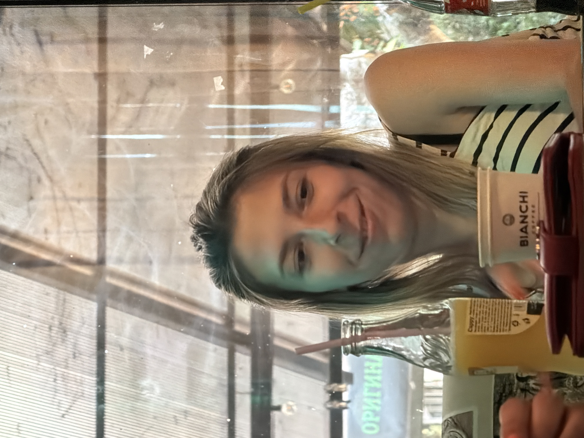 The image shows a young woman sitting at a table, possibly in a cafe or restaurant, with a bottle of orange juice and a cup. The setting appears to be indoors, near a window with a view of foliage outside. The woman has blonde hair and is smiling at the camera. A purse or wallet is visible on the table in front of her. The lighting suggests it is daytime. The text "ОРИГИН" is visible on a sign in the background through the window. The overall atmosphere is casual and relaxed. There is no specific indication of the city of Gulyantsi, Bulgaria within the image content.