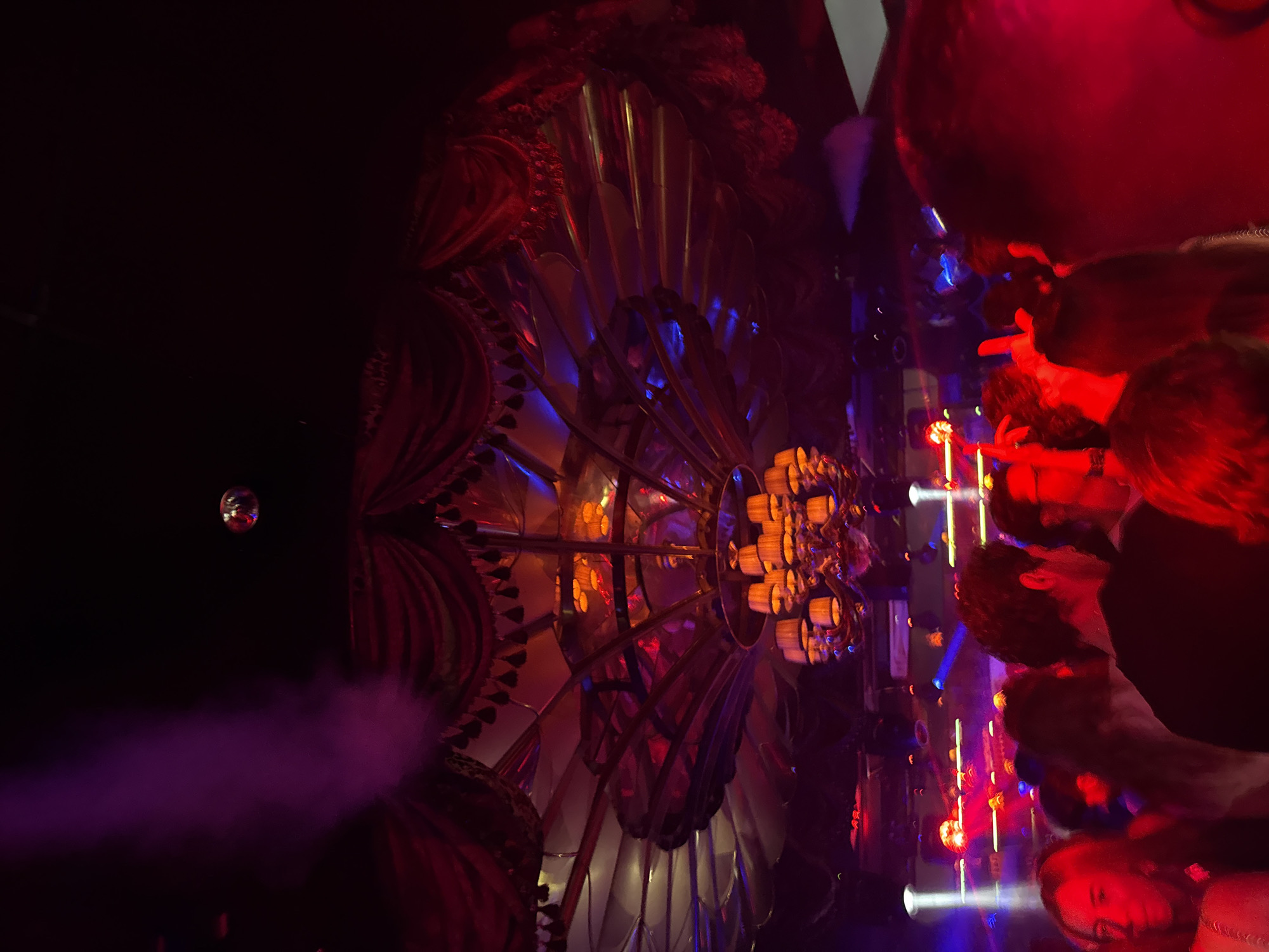 The image captures a dimly lit interior scene, likely within a venue in Mexico City, Mexico, given the ornate, stylized architecture. The focal point is a large, radial decorative structure with metallic, petal-like components, and a central arrangement of cylinder-shaped objects illuminated with orange light. The structure is adorned with red and blue lighting. In the foreground, indistinct figures of people are visible, bathed in red light. To the left, there is a disco ball hanging in the dark space, accompanied by purple smoke. The overall atmosphere suggests a club or event setting.