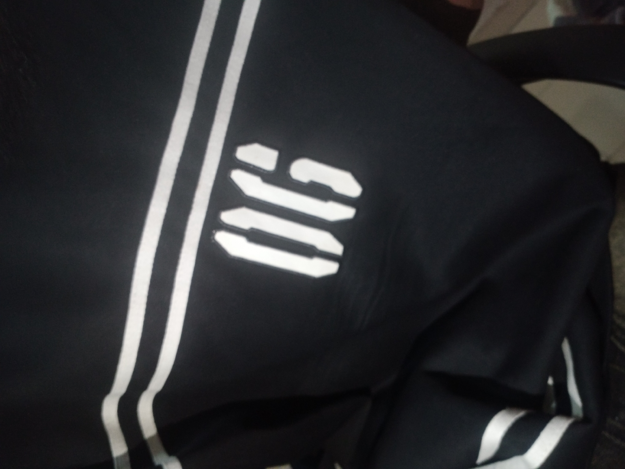 The image shows a close-up of a black garment, possibly athletic wear, with three white stripes running vertically down the left side. On the right side of the garment, the white numbers "06" are visible, appearing to be a stencil or embroidered design. The fabric appears to be synthetic and slightly creased. The background is blurred and out of focus, suggesting the garment is laid out or draped. There are no people or a discernible urban environment visible. The location in Jalingo, Nigeria is not determinable from the image content.