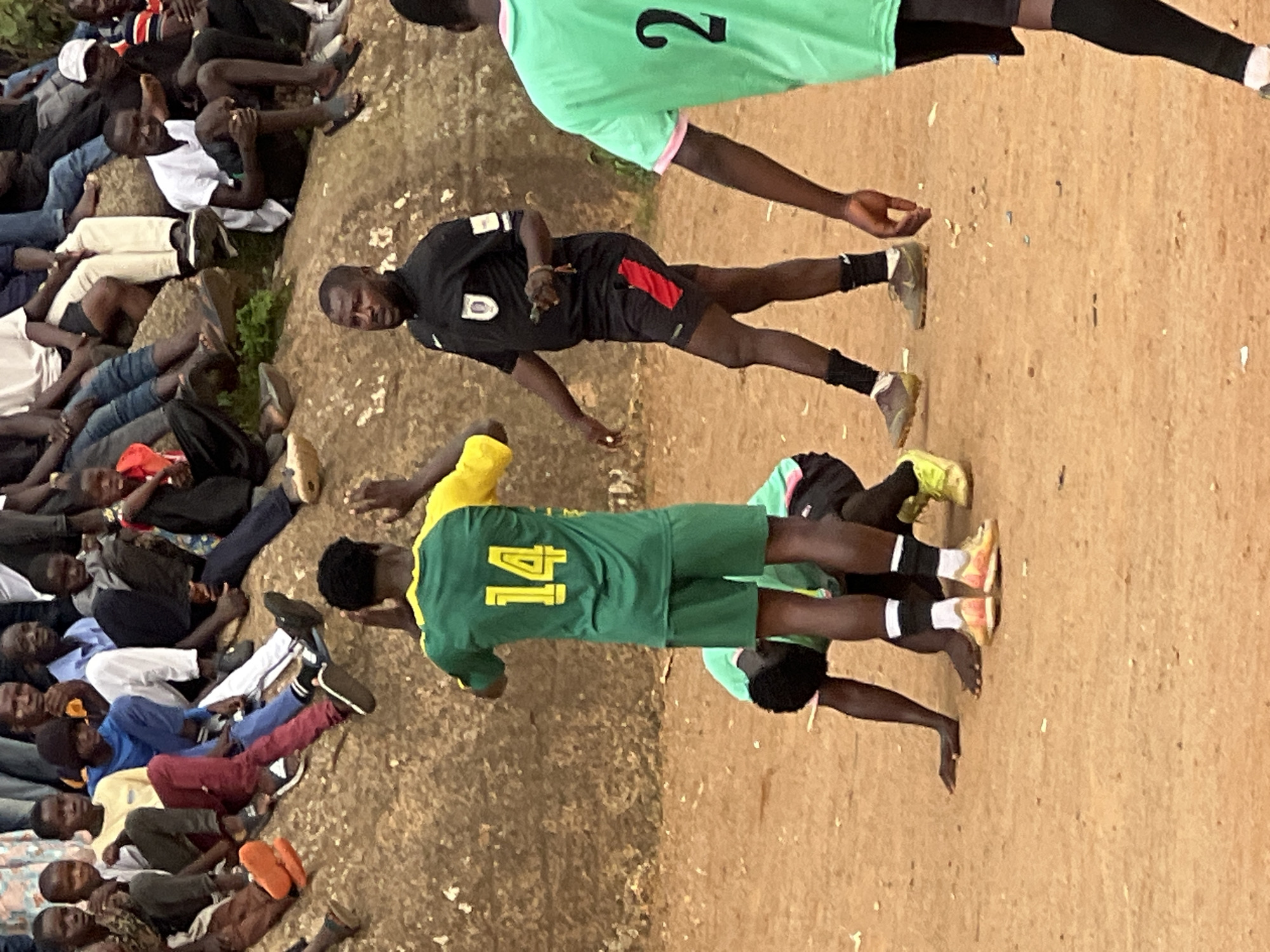 In the city of Jos, Nigeria, a group of young men are engaged in a soccer game on a dirt field. A referee, wearing a black shirt and shorts with red accents, stands in the center of the frame, holding a whistle and looking to his left. He appears to be observing the action on the field. To his right, a player in a green and yellow jersey with the number "14" on the back, faces away from the camera, his arms raised. Another player, wearing a green jersey, is on the ground in front of the referee, his body partially obscured. In the background, a crowd of spectators sits on a rocky embankment, watching the game. The overall atmosphere is one of casual sporting activity.