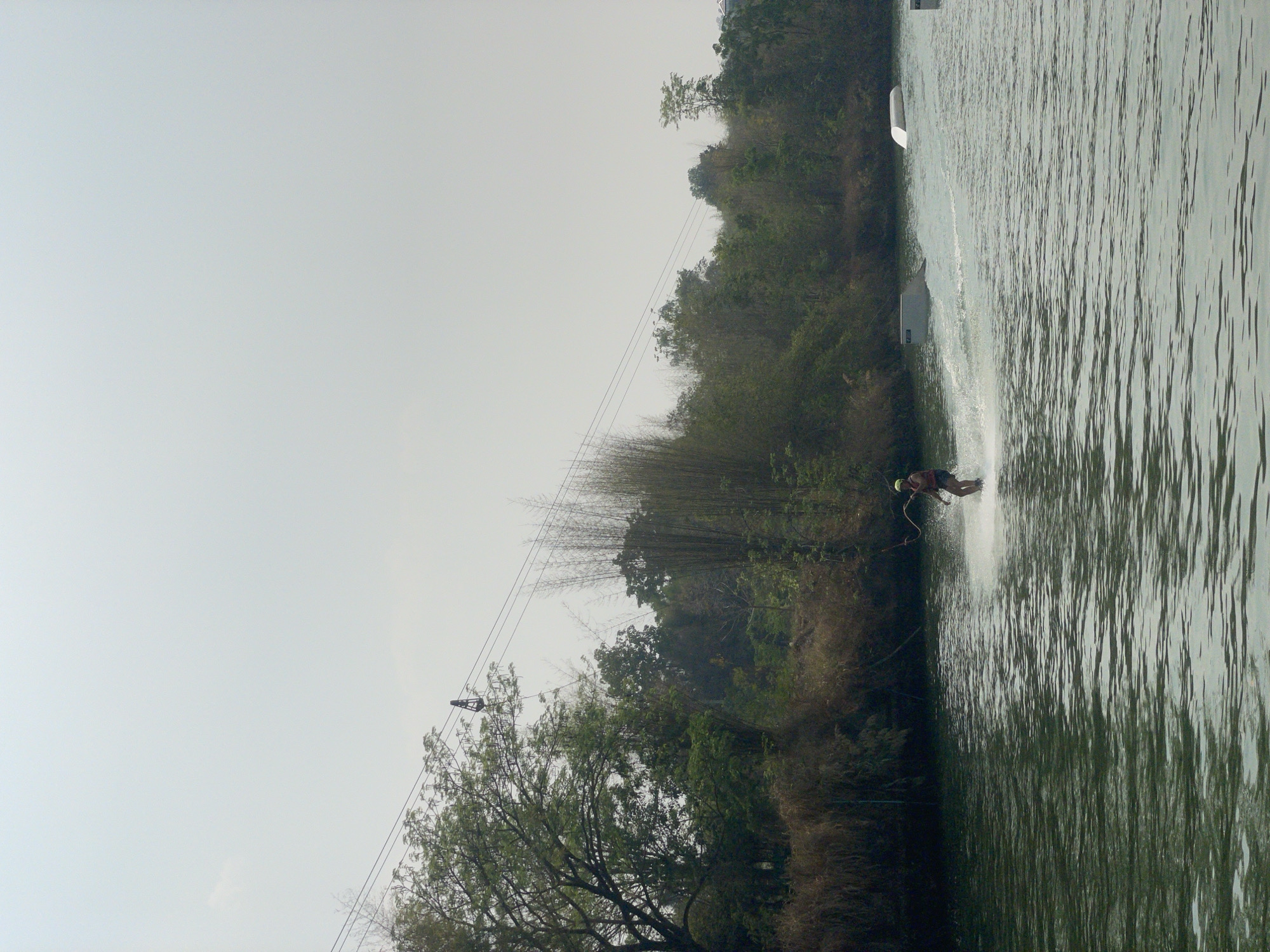 A vertical shot depicts a person water skiing in a lake, with the background featuring trees and a cloudy sky. The person is in motion, generating a spray of water. A cable system with a carrier is visible above the lake, likely used for pulling the skier. The scene is located in Tambon Nam Phrae, Thailand, as indicated by prior knowledge. A white floating object is visible in the water on the left side of the image.