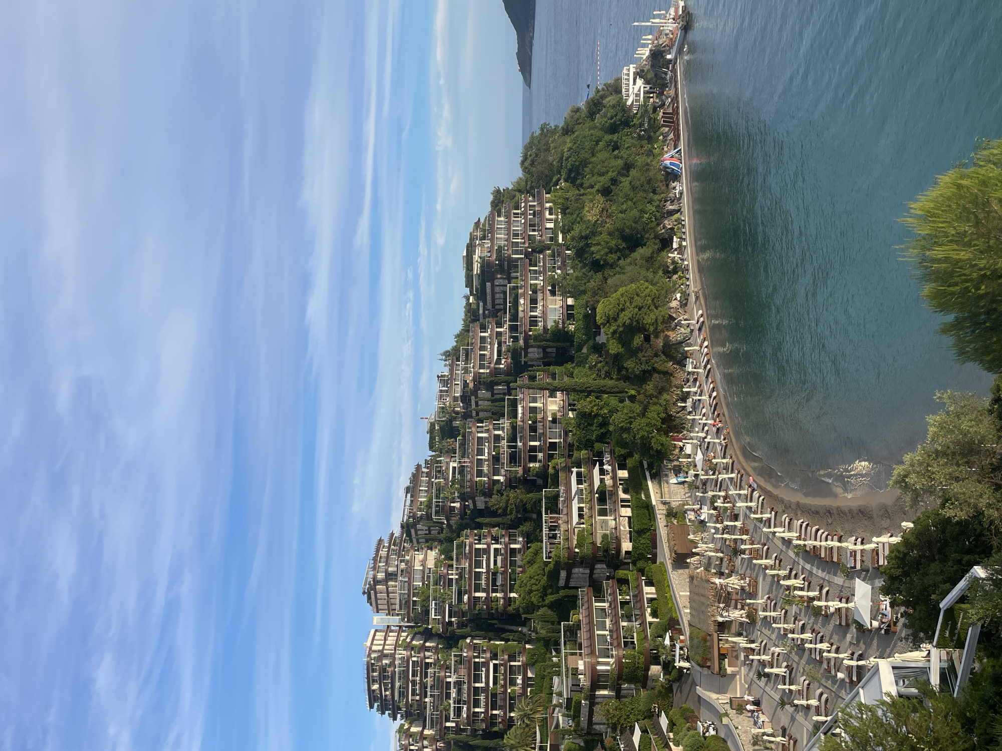 A high-angle shot captures a resort complex cascading down a lush green hillside towards a serene bay. The buildings, characterized by their terraced architecture, feature numerous balconies adorned with foliage. A sandy beach stretches along the coastline, lined with rows of empty lounge chairs and umbrellas. The clear, blue water of the Adriatic Sea gently laps at the shore. In the distance, a mountainous island can be seen rising from the sea under a partly cloudy sky. This scene depicts the luxurious waterfront of Budva, Montenegro, likely a part of the Sveti Stefan area.