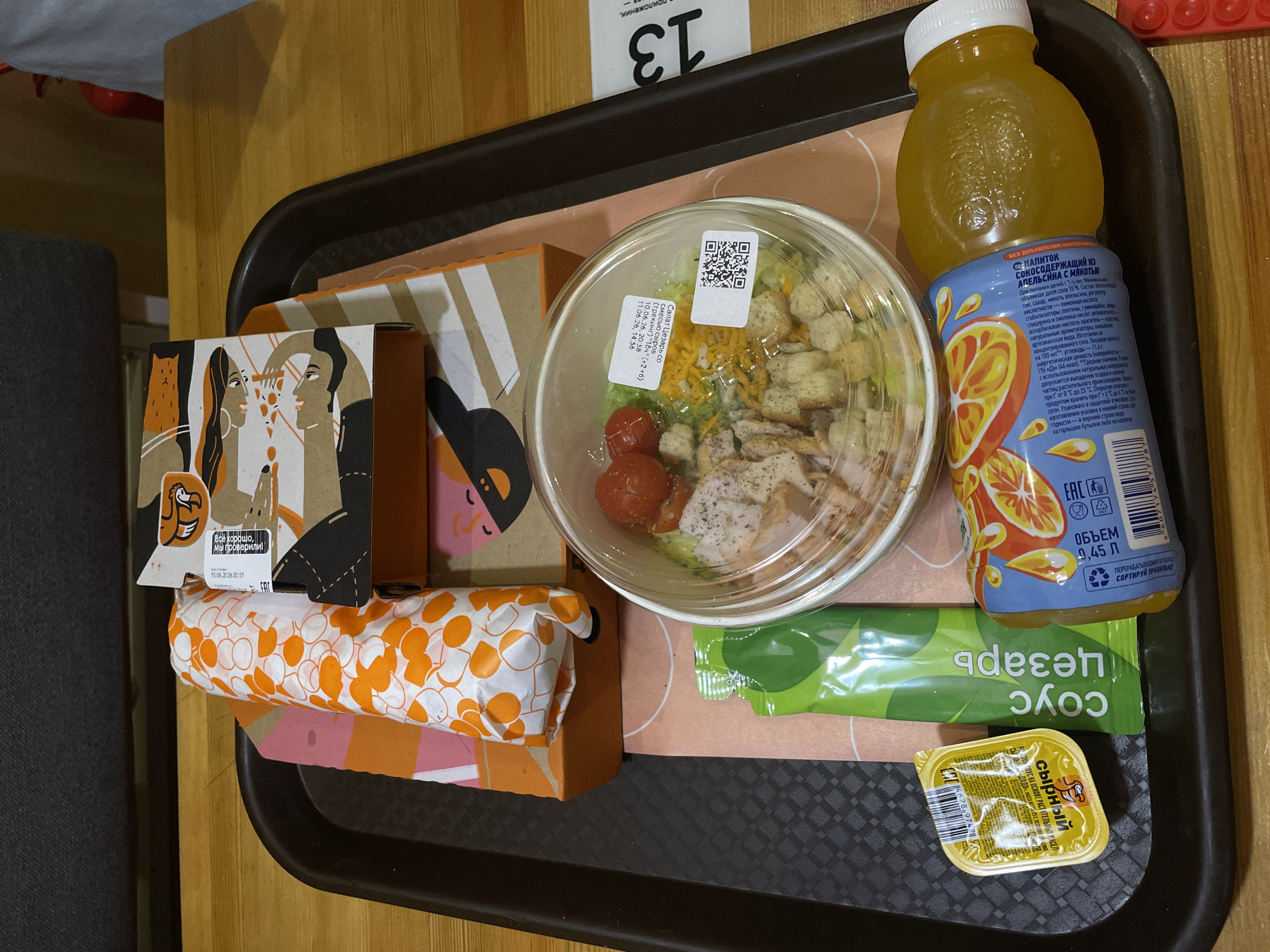 A fast-food meal is presented on a dark gray plastic tray. The tray contains a wrapped sandwich with an orange and white patterned wrapper, a brown cardboard box with a cartoon design on it, a clear plastic container of Caesar salad with chicken and croutons, a small yellow packet of sauce, a green packet of Caesar sauce, and a bottle of orange juice. The tray rests on a wooden table. A white card with the number "13" is visible to the right of the tray. The background is slightly out of focus but appears to be an indoor setting, possibly a fast-food restaurant. The meal seems to be ready to be consumed. The setting is Saint Petersburg, Russia.