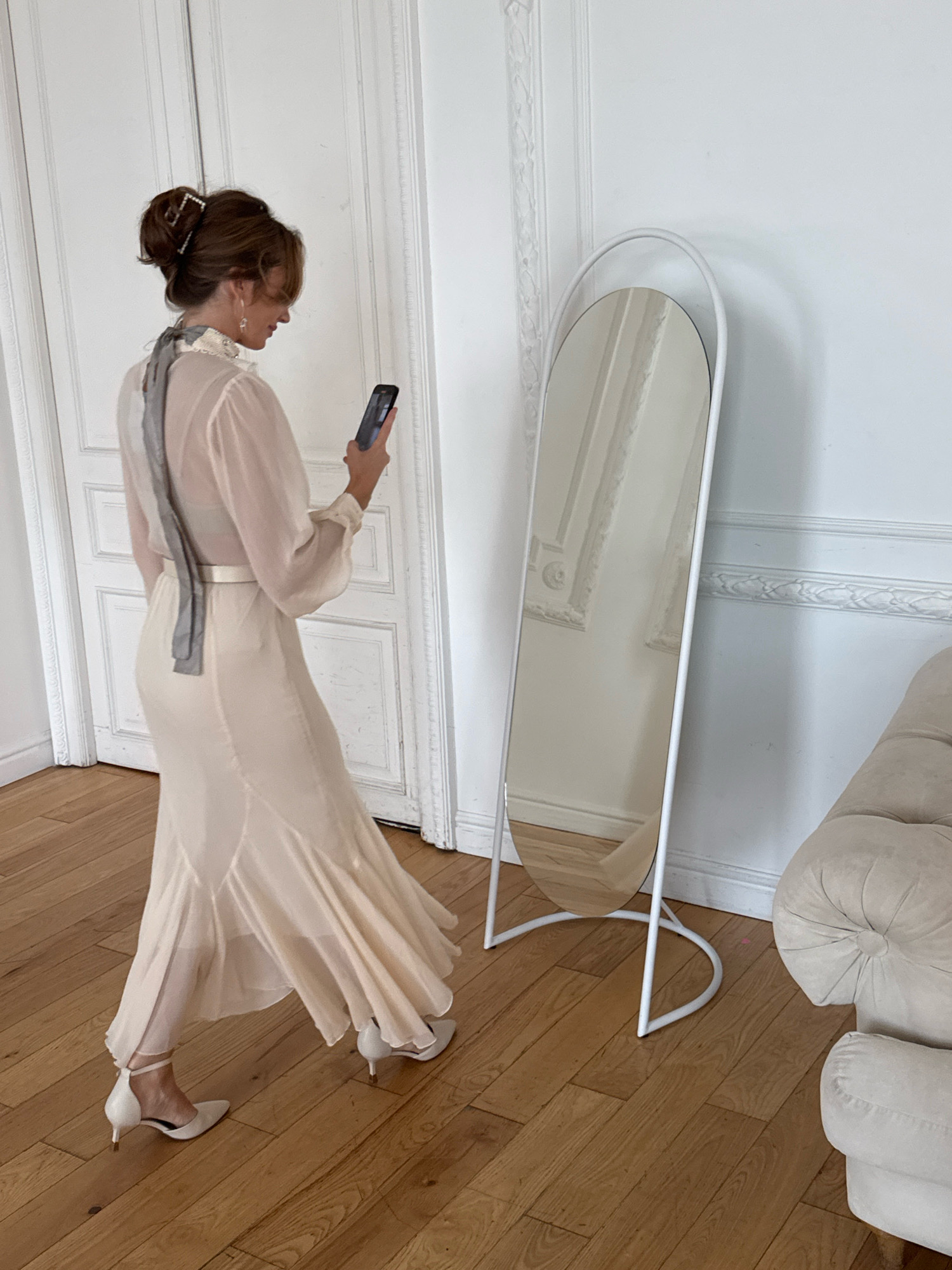 In Saint Petersburg, Russia, a woman stands in a room with light wood floors and white walls. She is facing away from the camera, looking at her phone, and is positioned near a tall, oval-shaped mirror with a white frame. She has brown hair styled in an updo with a decorative clip, and she is wearing a light-colored, sheer dress with a ruffled neckline, a belt, and a flowing skirt. A gray ribbon is tied around her neck. She is also wearing white pointed-toe heels. To the right of the mirror, a portion of a light-colored, tufted sofa is visible. The room features decorative white molding on the walls and a white double door in the background.