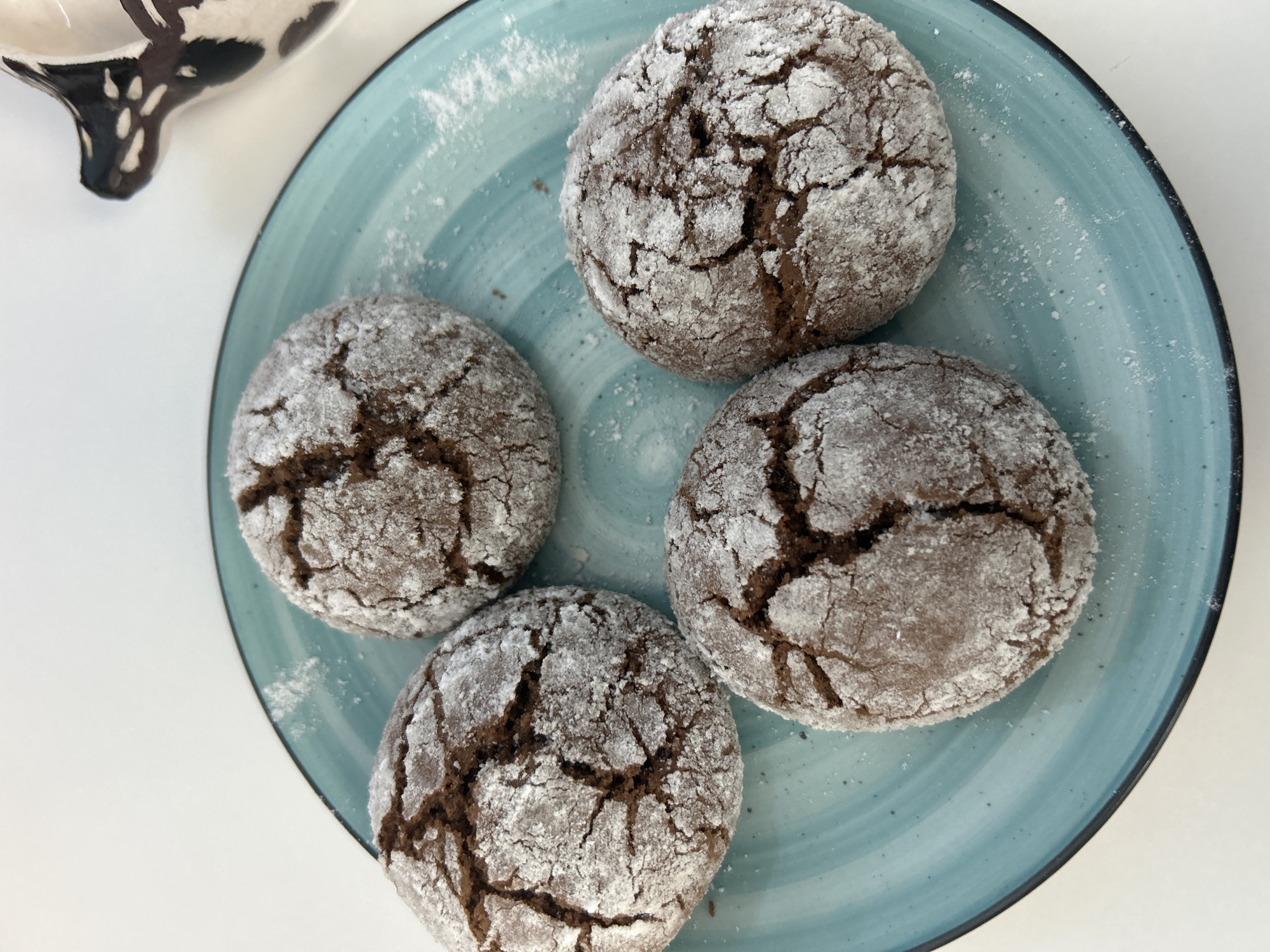 Four chocolate crinkle cookies, dusted with powdered sugar, are arranged on a teal-colored plate. The plate has a swirling pattern and a dark rim. A white surface is visible around the plate, and a portion of a black and white patterned object, possibly a pitcher or creamer, is in the upper right corner. The image is a close-up shot, and no specific location in Orenburg, Russia, can be determined from the visual information provided.