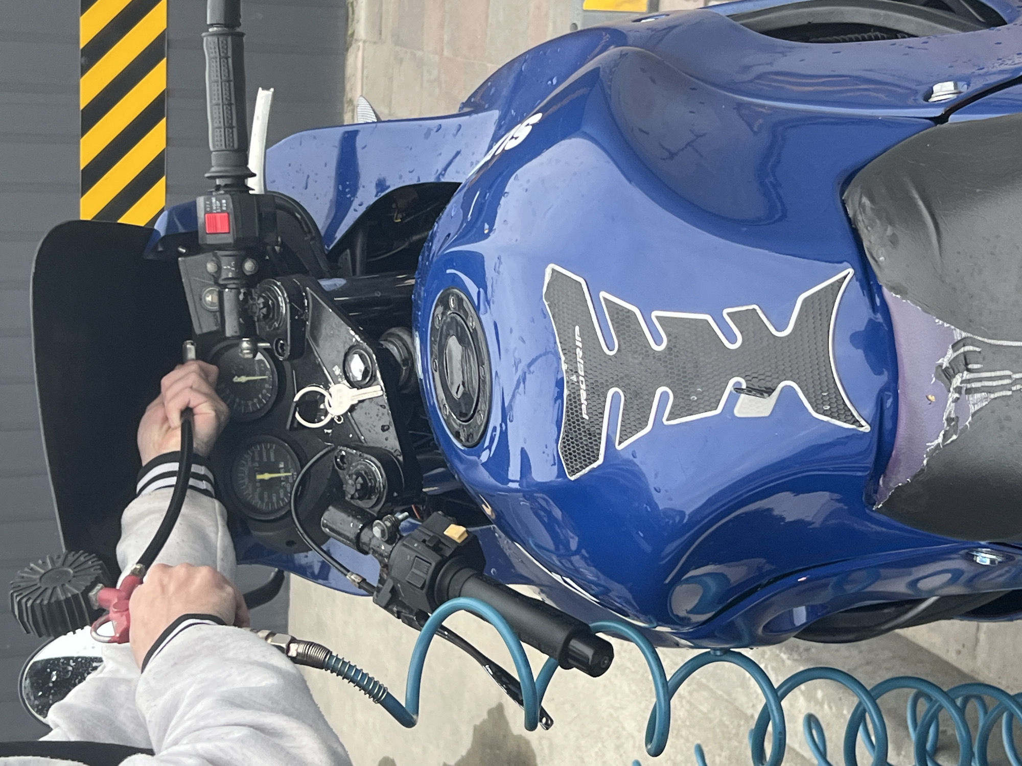 A blue motorcycle is being washed by a person in a grey hoodie. The motorcycle has a black-and-white graphic on its side. The person is using an air compressor with a blue hose to blow dry the instrument panel. The background shows a grey building with yellow and black warning stripes. The location is Horodok, Ukraine.