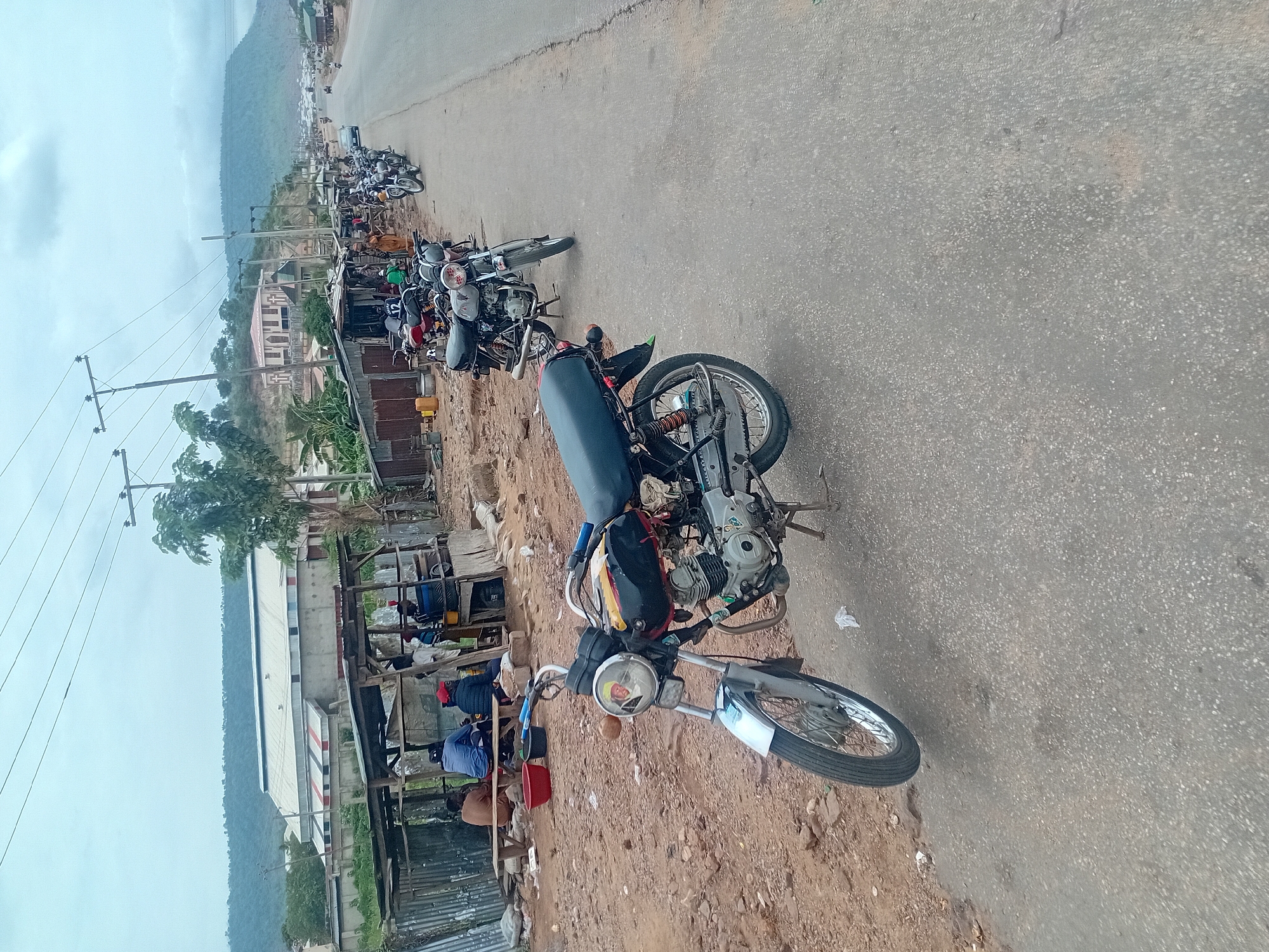 The image shows a street scene in an unidentified location. The perspective is from ground level, slightly angled upward. The primary subject is a line of motorcycles parked along the side of a paved road. The nearest motorcycle is a close-up view. Beyond the row of motorcycles, a makeshift building or stall is visible along with other structures and people in the background. In the far distance, a hill or mountain range and overcast sky are seen. Utility poles with wires run along the roadside. The ground adjacent to the road is dirt and gravel.