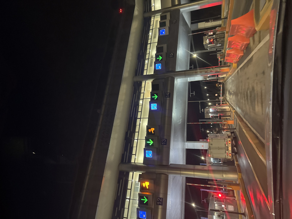 This image depicts a nighttime view of a multi-lane toll plaza in Ocoyoacac, Mexico, captured from the interior of a vehicle approaching the facility. The scene is illuminated by artificial lights, with the pavement appearing wet and reflective.
The central subject is a modern toll plaza structure with a broad, flat roof supported by numerous cylindrical pillars. This overhead structure features rows of illuminated panels or windows. Below the structure, at least five distinct toll lanes are visible, numbered 12 through 16 from left to right. Each lane is equipped with an overhead digital sign system displaying combinations of a blue square with a white dash (indicating electronic payment), a green downward arrow (indicating an open lane), and an orange hand symbol (indicating manual payment or cashier service). For example, lanes 12 and 13 show all three symbols, while lanes 14 and 15 show the blue square and green arrow.
Within the lanes, physical toll booths or kiosks are present, along with automated red and white striped traffic barriers, some of which are lowered (e.g., Lane 12). Red traffic signals are visible at various lanes, including Lane 12. A white box truck, with its rear lights illuminated, is positioned in one of the central lanes, appearing to be stationary or moving slowly through a toll booth. The far right lane displays orange plastic road barriers. No individuals are clearly visible within the scene. The background beyond the toll plaza shows additional streetlights under a dark night sky.