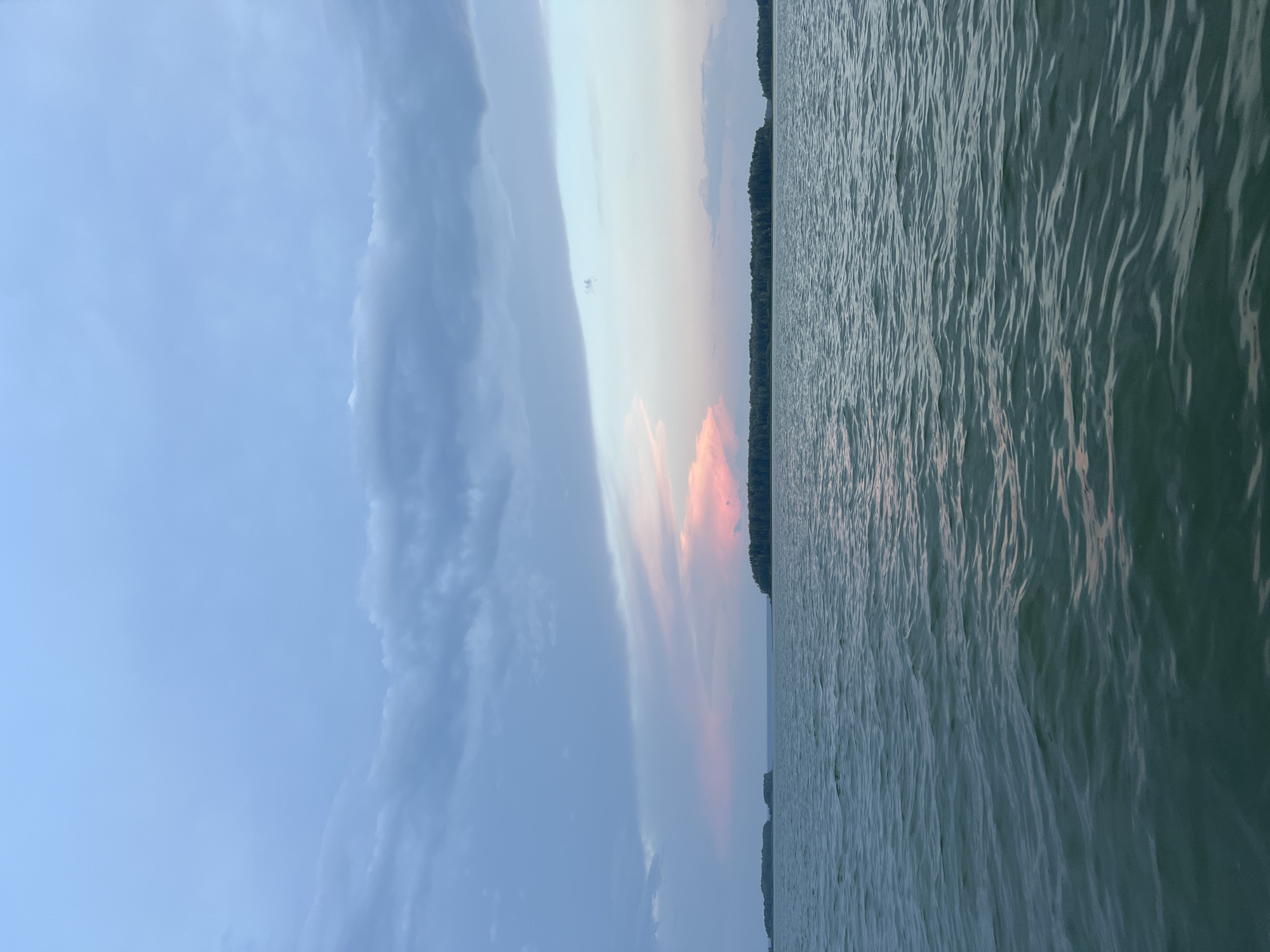 The image depicts a tranquil body of water, likely a lake or a wide river, under a dusky sky. The water is a deep green hue with gentle ripples, reflecting the muted colors of the sky. In the distance, a long, tree-lined shoreline stretches across the horizon, interrupted by a few small, sparsely vegetated islands. The sky is a gradient of blues and grays, with hints of soft pink and orange from a setting sun, suggesting the time of day is late evening. The overall atmosphere is serene and natural, devoid of any human presence or man-made structures. The location is identified as Novopichugovo, Russia.