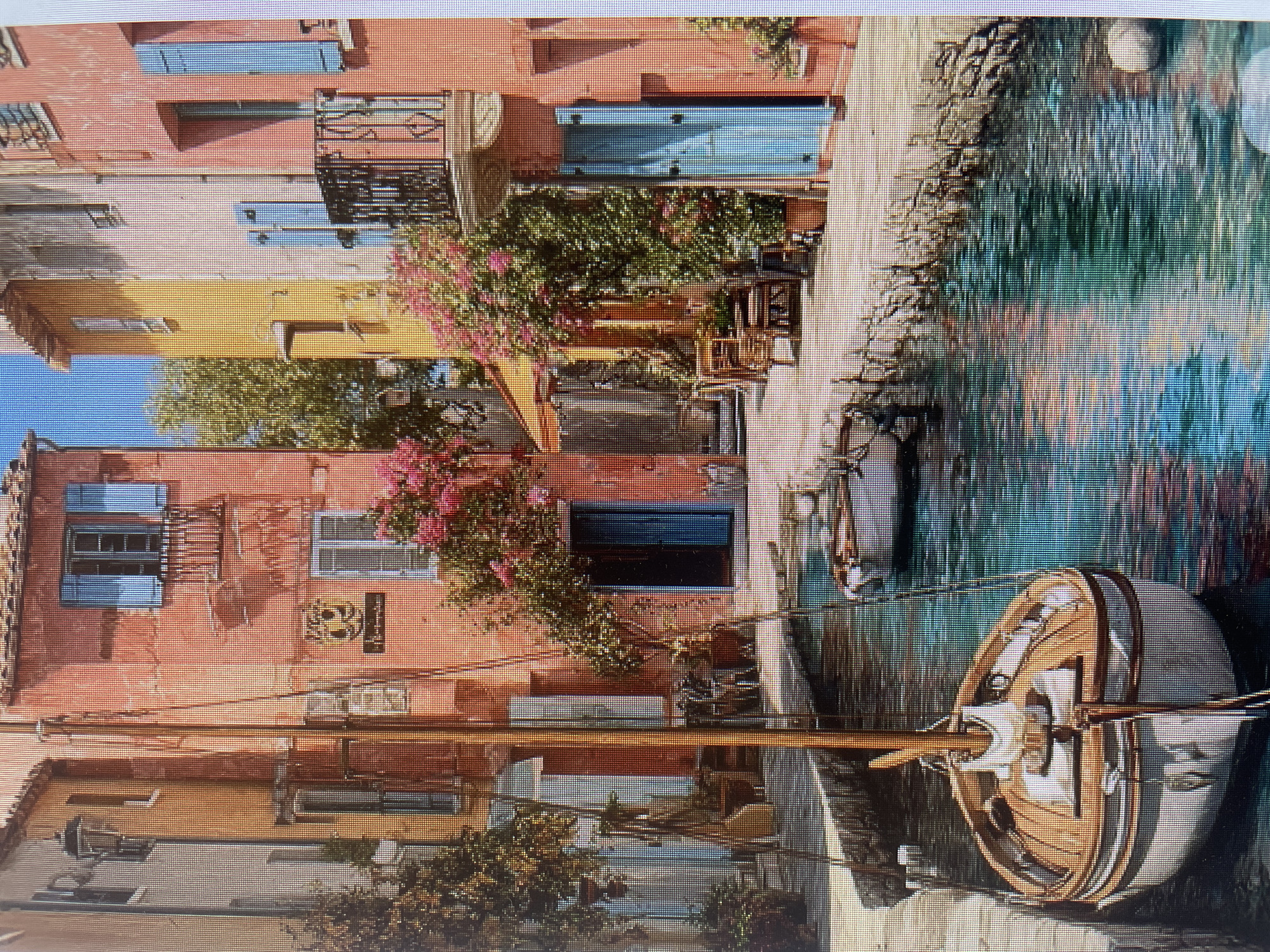 The image displays a picturesque, colorful canal-side village or town scene under clear blue skies. Multi-story buildings with pastel-colored facades, predominantly terracotta red/pink and ochre yellow, line the left bank of a narrow waterway.
The buildings feature numerous windows, many adorned with bright blue or white shutters, some open, some closed. Several structures include black wrought-iron balconies with ornate railings. Lush green foliage and vibrant pink/magenta flowering plants, possibly bougainvillea, spill from balconies and climb the building walls. Green trees are visible between and behind some of the structures.
A tranquil, green-blue canal occupies the right side of the frame, reflecting the buildings and the sky. Several boats are moored in the water. In the immediate foreground on the right, a classic wooden sailboat with a light-colored hull, wooden decking, a mast, and visible rigging is prominently featured, with its reflection visible in the water.
Small details include outdoor seating areas with tables and chairs near some buildings, partially obscured by greenery. On one of the reddish buildings, a dark sign with a yellow emblem and some illegible text is visible, as well as a white sign below it. The scene appears to be set during a sunny daytime, with distinct shadows indicating bright natural light. No people are visible.