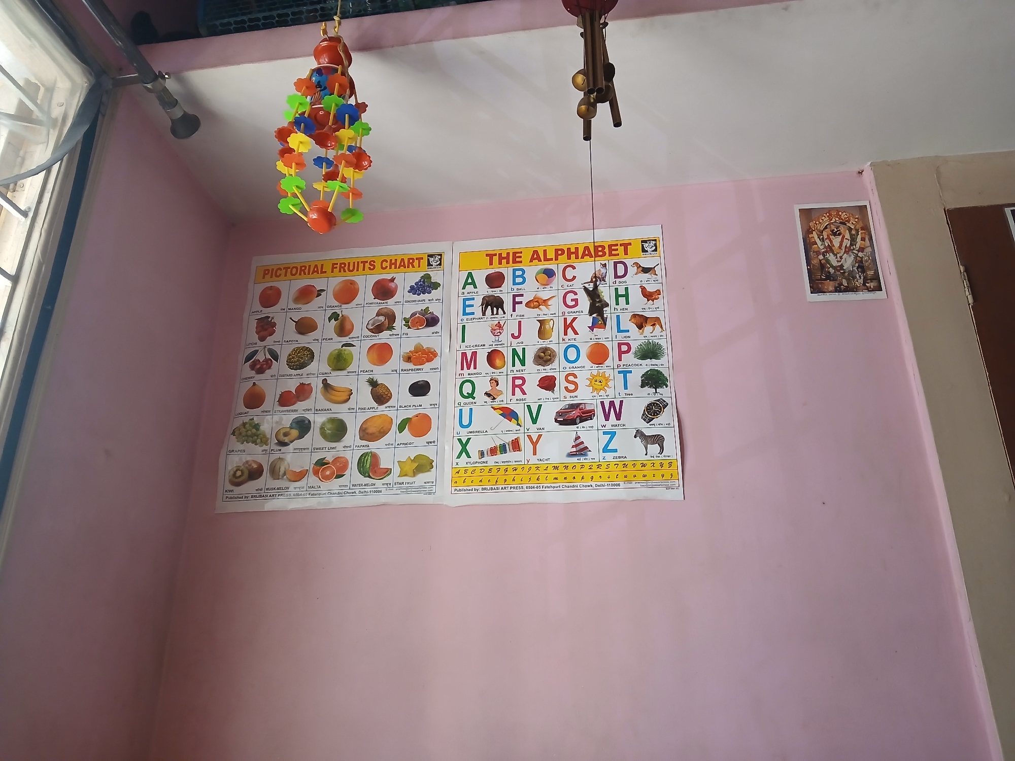 A low-angle indoor shot shows a pink wall decorated with two educational charts and a framed picture. The chart on the left is titled "PICTORIAL FRUITS CHART" and the chart on the right is titled "THE ALPHABET." A framed picture is mounted on the wall to the right of the alphabet chart. A colorful hanging toy is suspended from the ceiling on the left and a wind chime hangs from the ceiling above the right chart. The location is likely a residence or classroom in India, based on the educational materials and decor. The street name of Fatehpuri Chandni Chowk printed on the charts confirms the location is NOT in the city of Bengaluru, India.