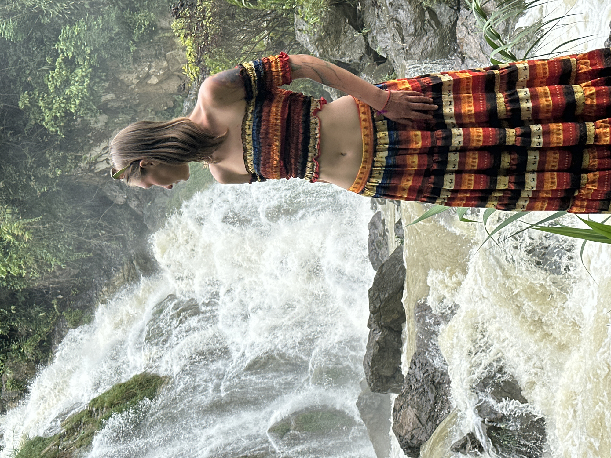 A person stands in front of a cascading waterfall in Bản Phúc, Vietnam. The person is wearing a colorful, off-the-shoulder crop top and a matching tiered skirt. The waterfall flows over rocks, creating white foam and spray. Lush green vegetation surrounds the waterfall and the person. The person is looking to the side, away from the camera, with sunglasses pushed up on their head. The overall scene depicts a natural, tropical environment.