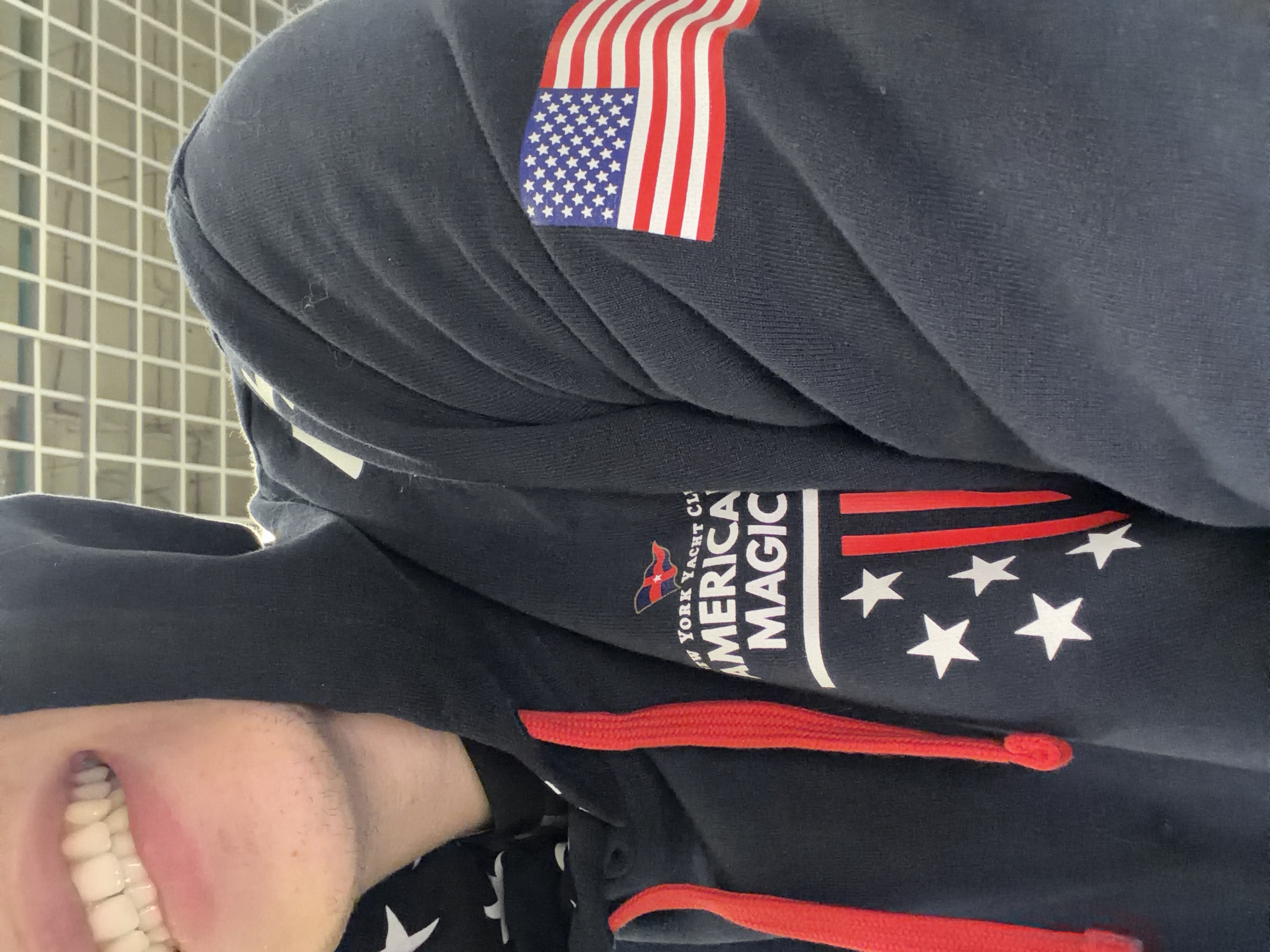 The image shows a close-up view of a person wearing a navy blue hoodie. The hoodie has red drawstrings and features an American flag graphic on the right sleeve. On the left side of the chest, there is a logo that reads "NEW YORK YACHT CLUB AMERICA MAGIC" above a design of stars and red stripes. A portion of the person's face is visible, showing their teeth and chin, which has light stubble. The background appears to be an indoor environment with a white grid ceiling. There is no discernible information about the location in Nizhny Novgorod, Russia.