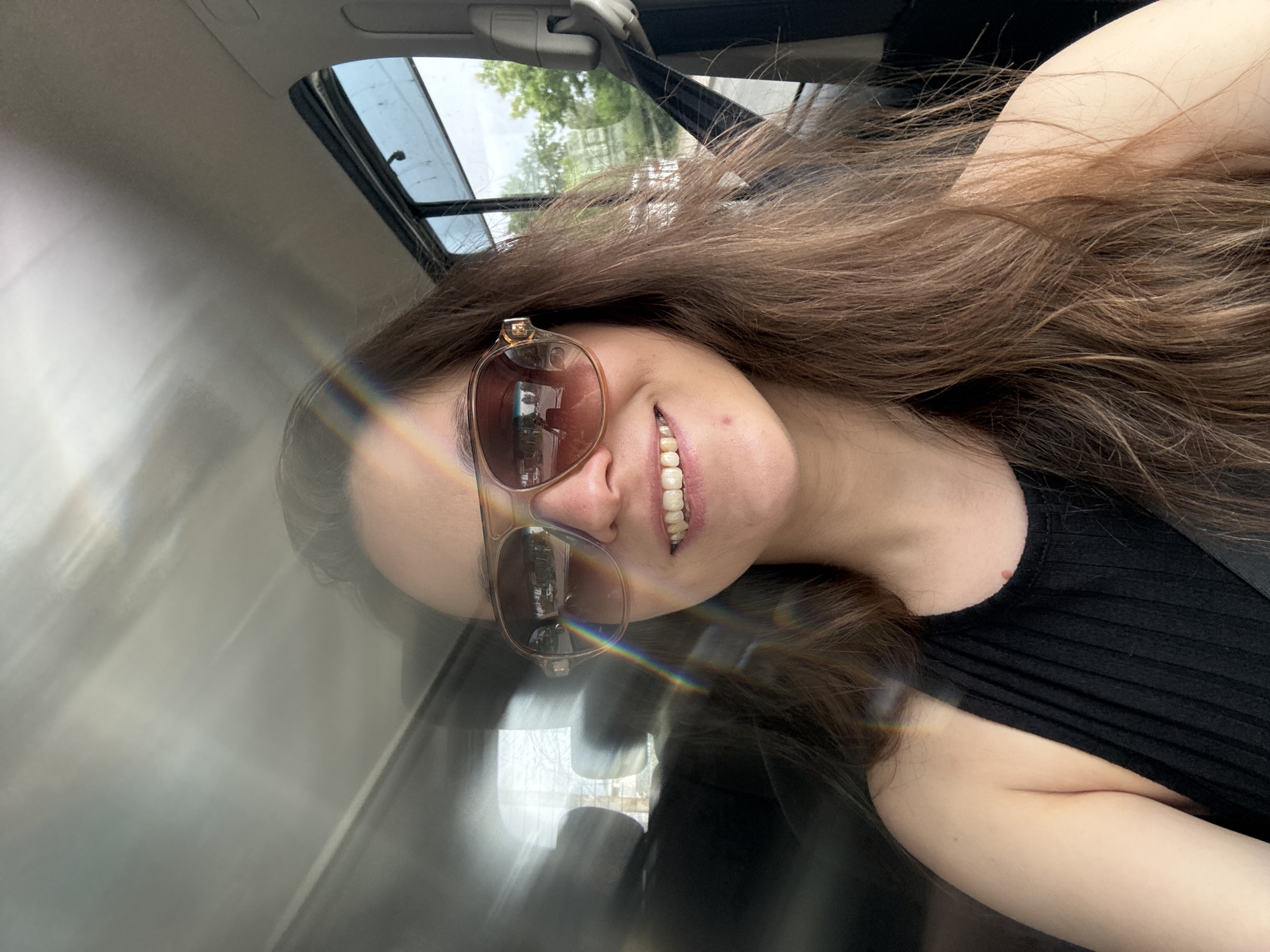 A medium shot depicts a woman with long, light brown hair wearing sunglasses and a black ribbed top, angled in what appears to be the driver's seat of a car. A rainbow reflection is visible on the car's interior. The car has a sunroof. The location is Chaikovsky, Russia.