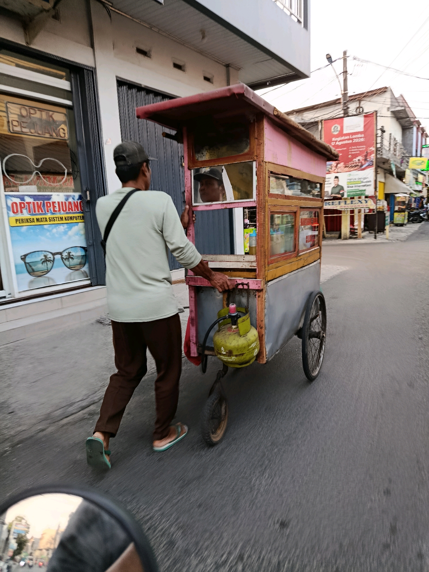 The image depicts a street scene in Bekasi, Indonesia. In the foreground, a man wearing a light green long-sleeved shirt, dark brown pants, a dark baseball cap, and flip-flops is pushing a food cart. The cart is made of wood and metal, painted pink and red, and has a glass display case. A green gas cylinder is attached to the cart. The man is walking on the right side of the street, moving away from the viewer. To the left of the man, there is a storefront with a sign that reads "OPTIK PEJUANG" and displays an advertisement for sunglasses. In the background, buildings line the street, including a building with a large red banner. Traffic lights and utility poles are visible. The street is paved and appears to be a commercial area. The reflection of traffic and buildings is visible in a motorcycle's rearview mirror in the bottom left corner of the image.